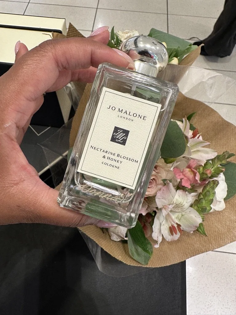 A hand holding a Jo Malone London cologne bottle labeled 'Nectarine Blossom & Honey', with a bouquet of flowers in brown wrapping paper in the background.