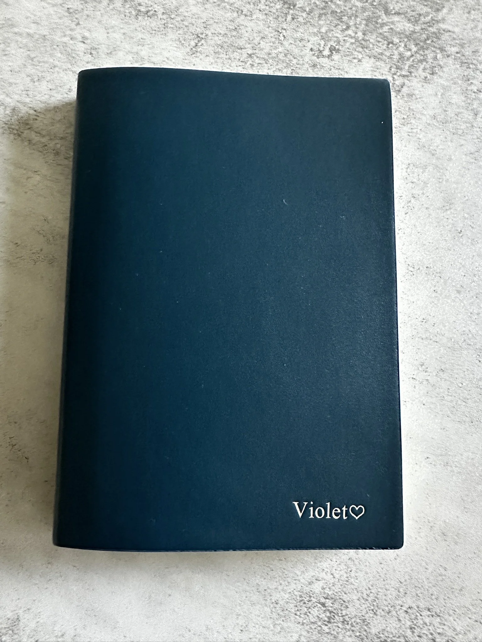 A closed, dark blue notebook with the name 'Violet' and a heart symbol printed in white in the bottom right corner, placed on a light gray textured surface.