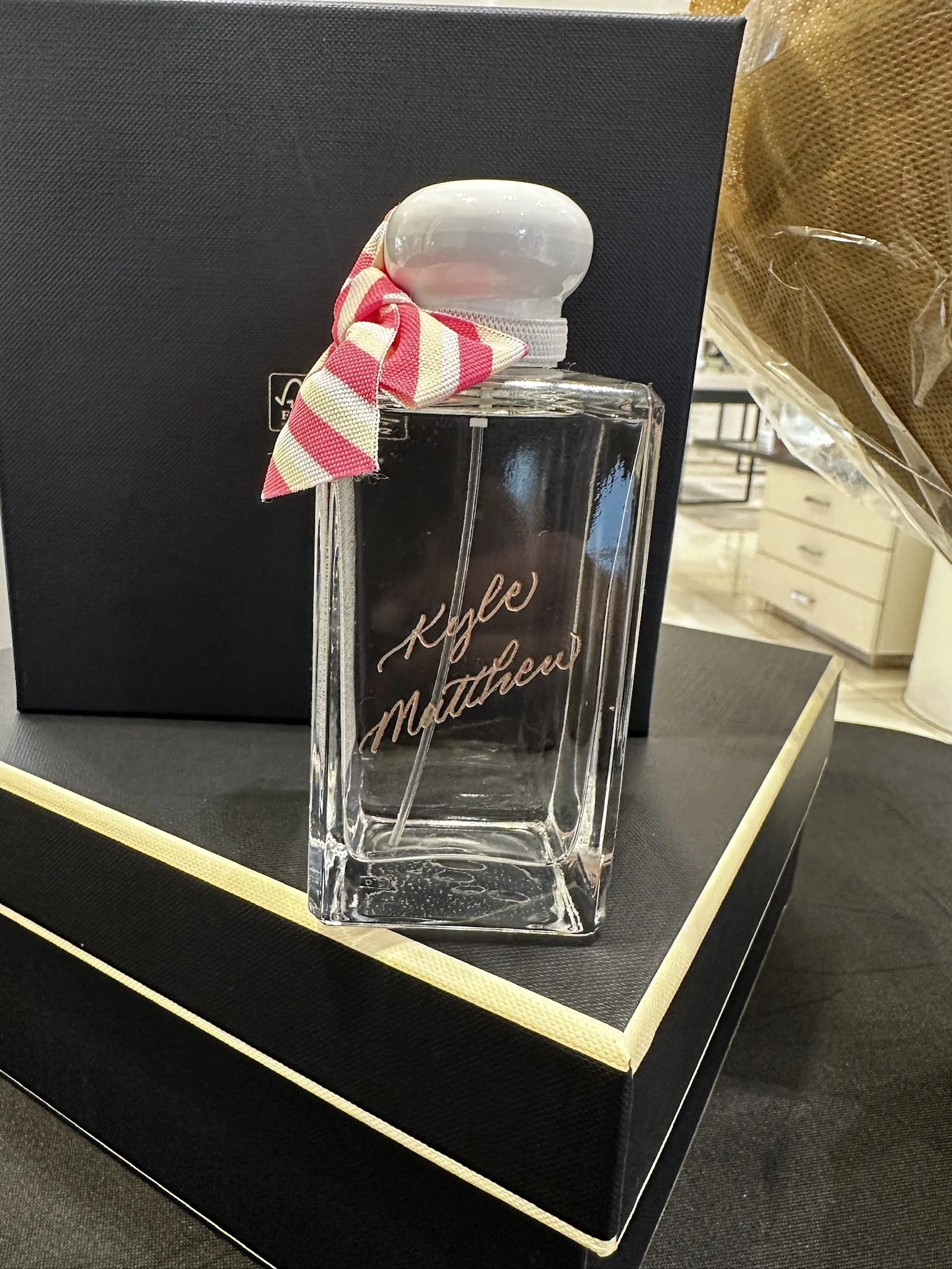 A clear glass perfume bottle with gold calligraphy writing, placed on a display platform with a black background and gold trim. 