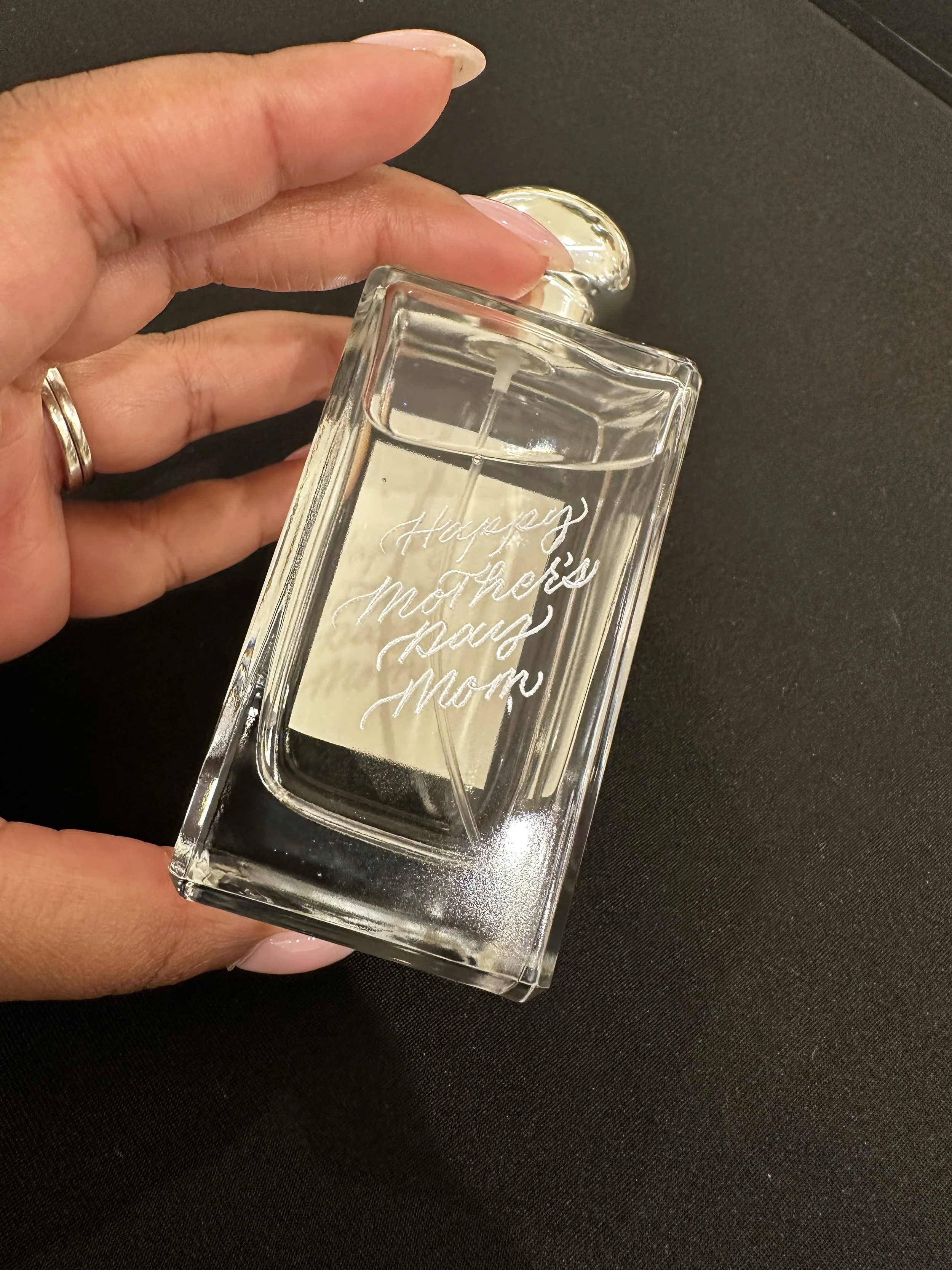 A hand holding a Jo Malone bottle of perfume. "Happy Mother's Day Mom" engraved with silver fill on a black background.