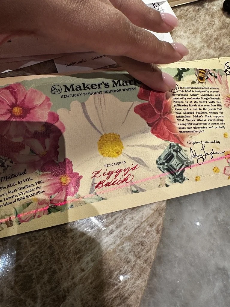 A person holding a paper label or certificate with text and colorful floral designs, including pink and red flowers, and a small bee illustration.