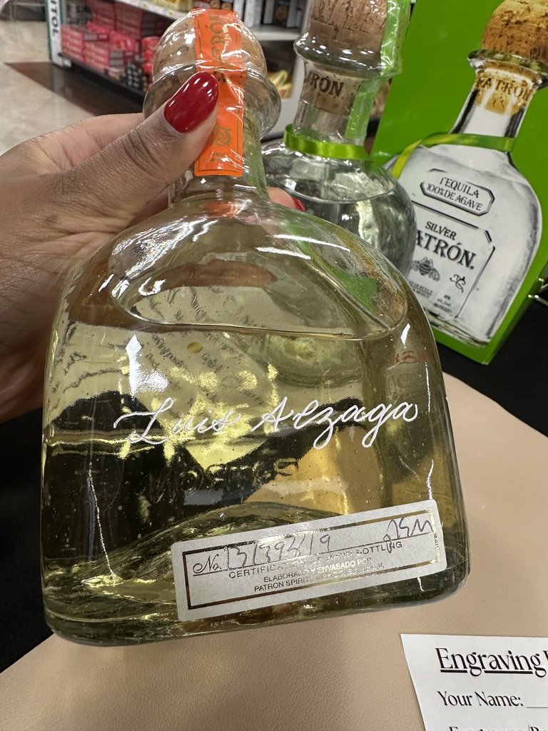 A hand with red nail polish holding a clear glass bottle with a light-colored liquid inside, labeled with the name 'Luz Alzaga'. In the background, there are other bottles on a store shelf, including a tequila bottle and a green box.