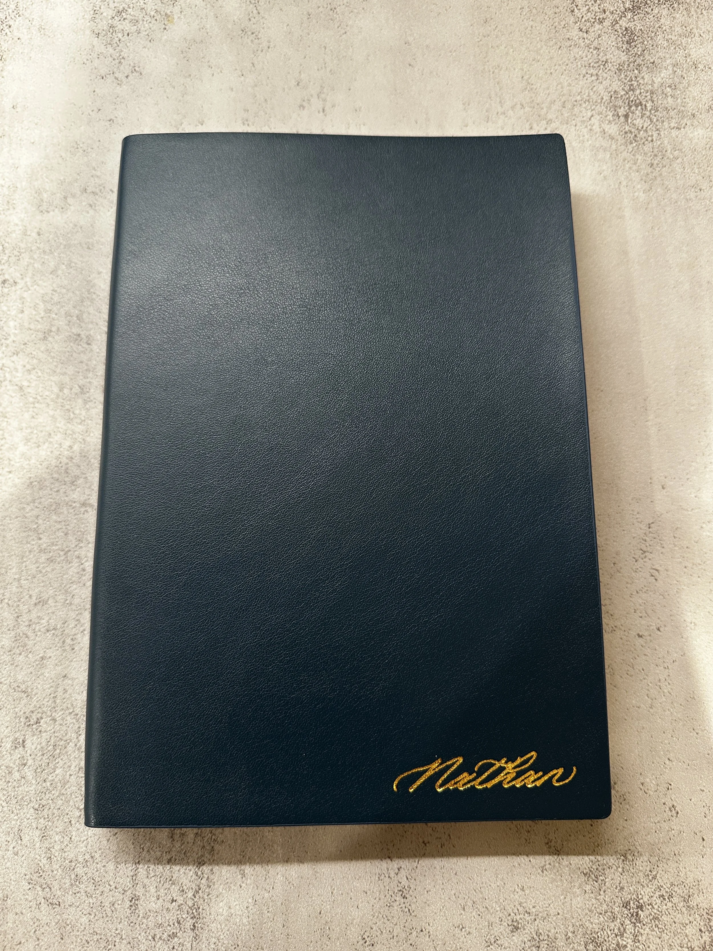 A closed, dark blue notebook with the name 'Nathan' in the bottom right corner in gold calligraphy, placed on a light gray textured surface.