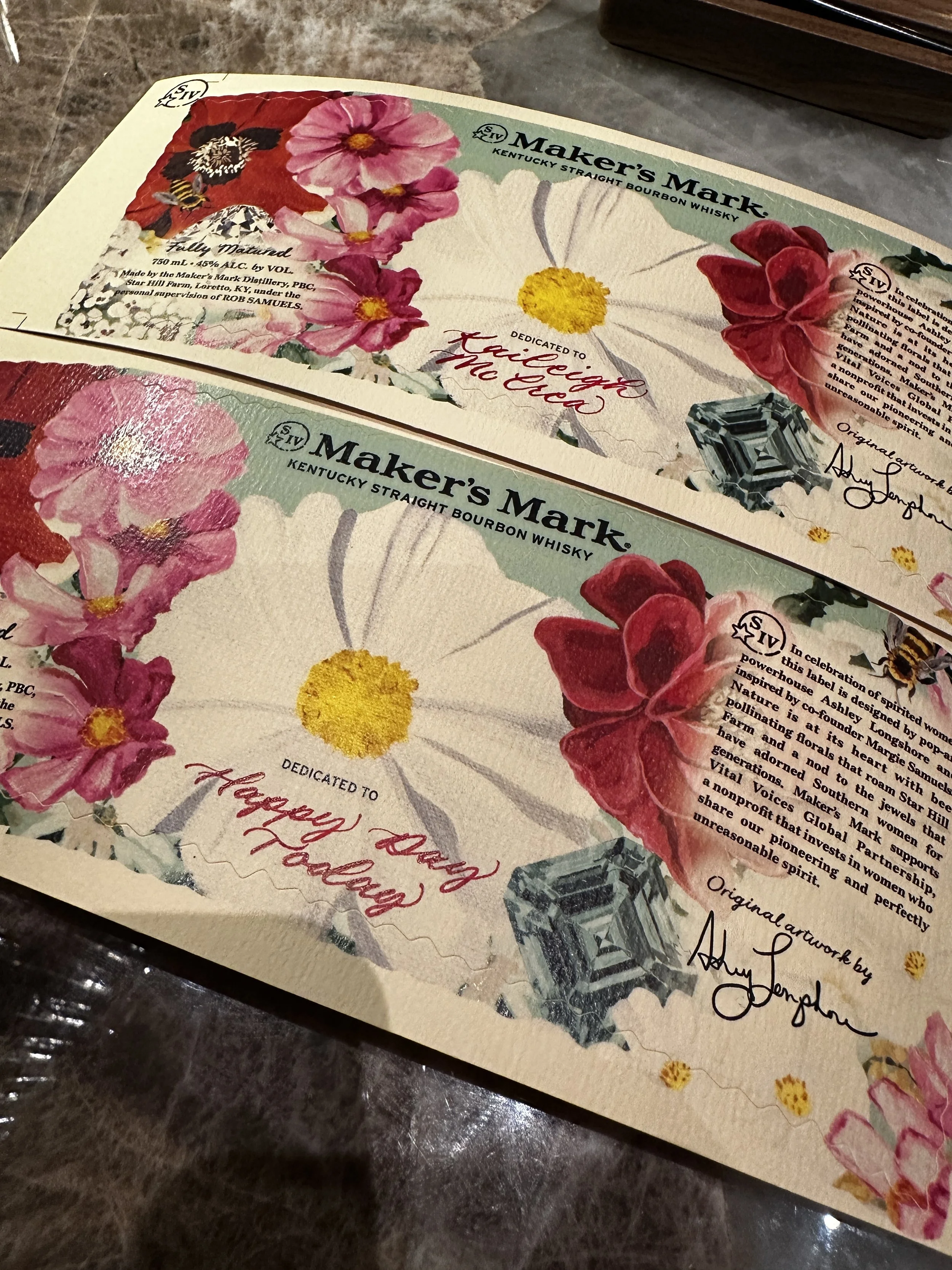 Close-up of a whiskey label with floral and bee illustrations, titled 'Maker's Mark Kentucky Straight Bourbon Whisky,' dedicated to 'Kaleigh and Joce,' signed by Ashley Johnplum, on a wooden surface.