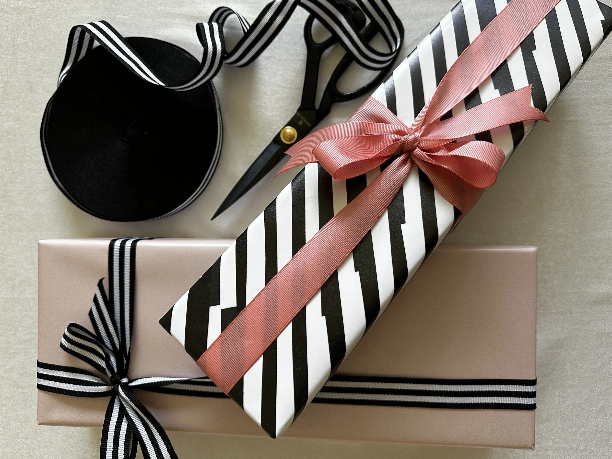 Two wrapped gift boxes with ribbons, a pair of scissors, and a roll of black and white striped ribbon on a light-colored surface. One box is pink with a black and white striped ribbon, and the other has black and white stripes with a pink ribbon.