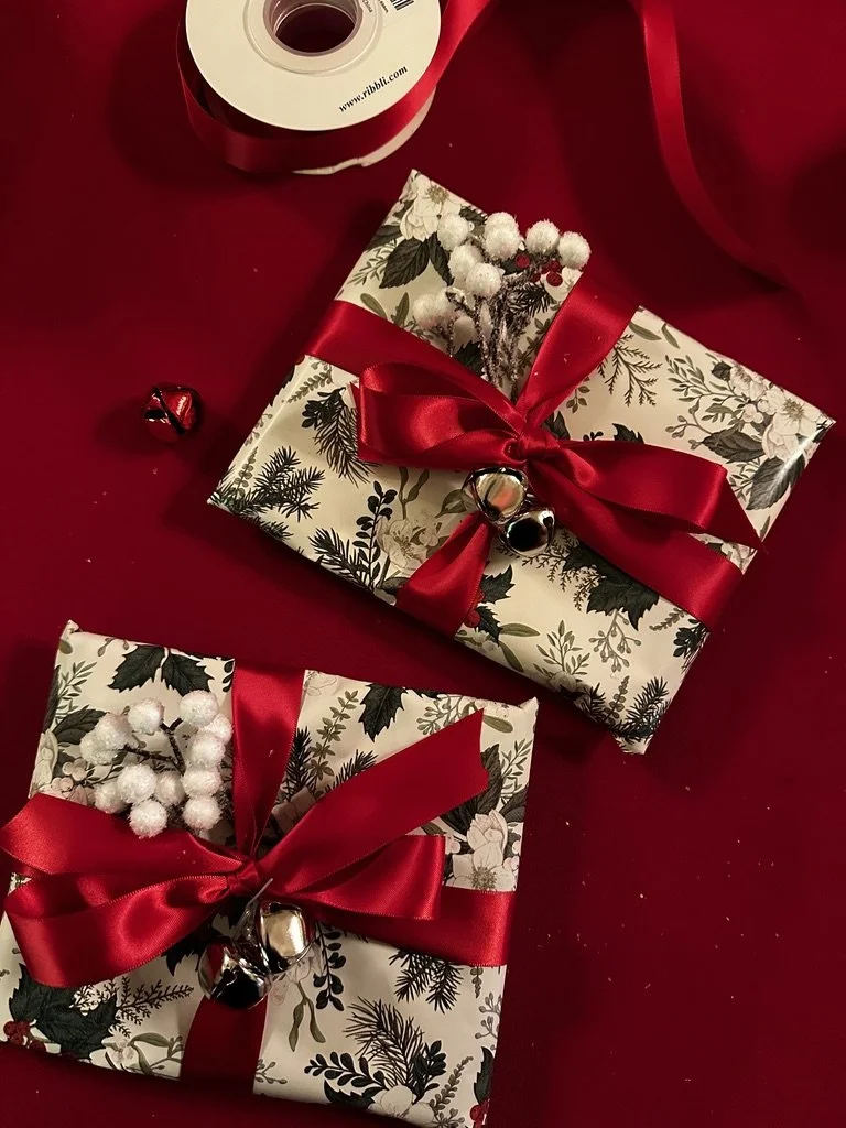Two Christmas presents wrapped in floral paper with holly and mistletoe designs, tied with red satin ribbons, on a red background, with small bells and a roll of ribbon nearby.