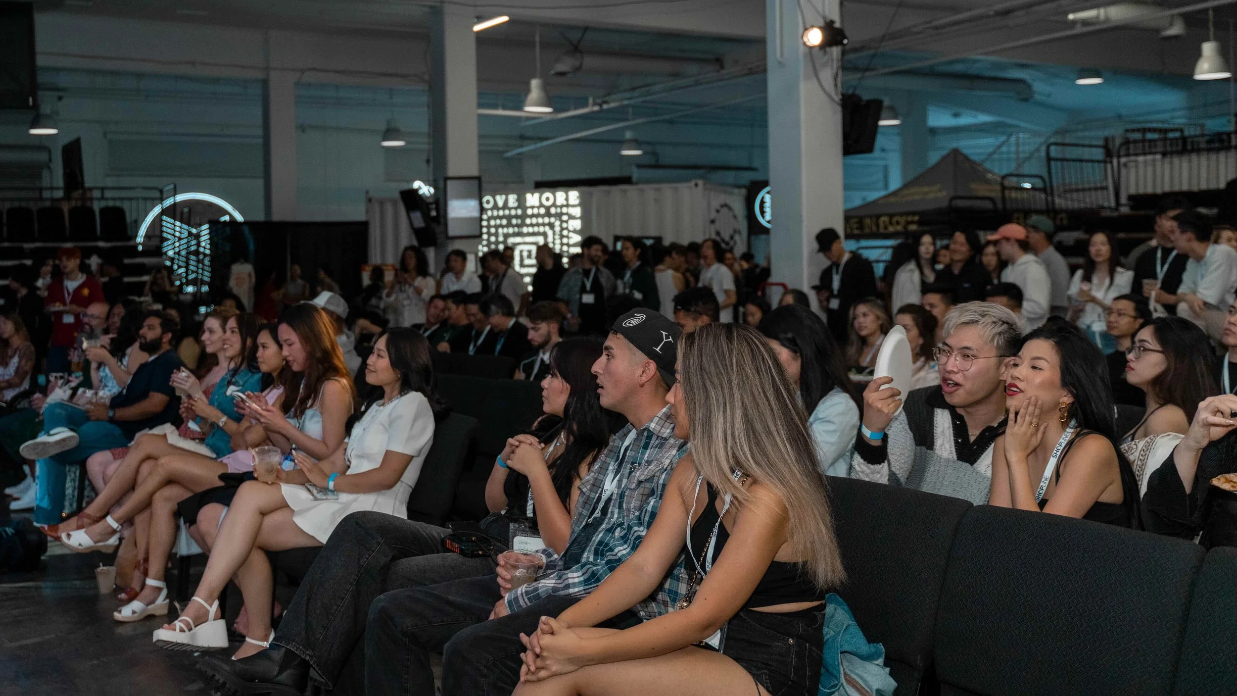 An audience at a conference or event, sitting in rows and engaging with the presentation, with some using phones and others conversing. The setting appears to be an indoor venue with dim lighting and visible stage or display area.