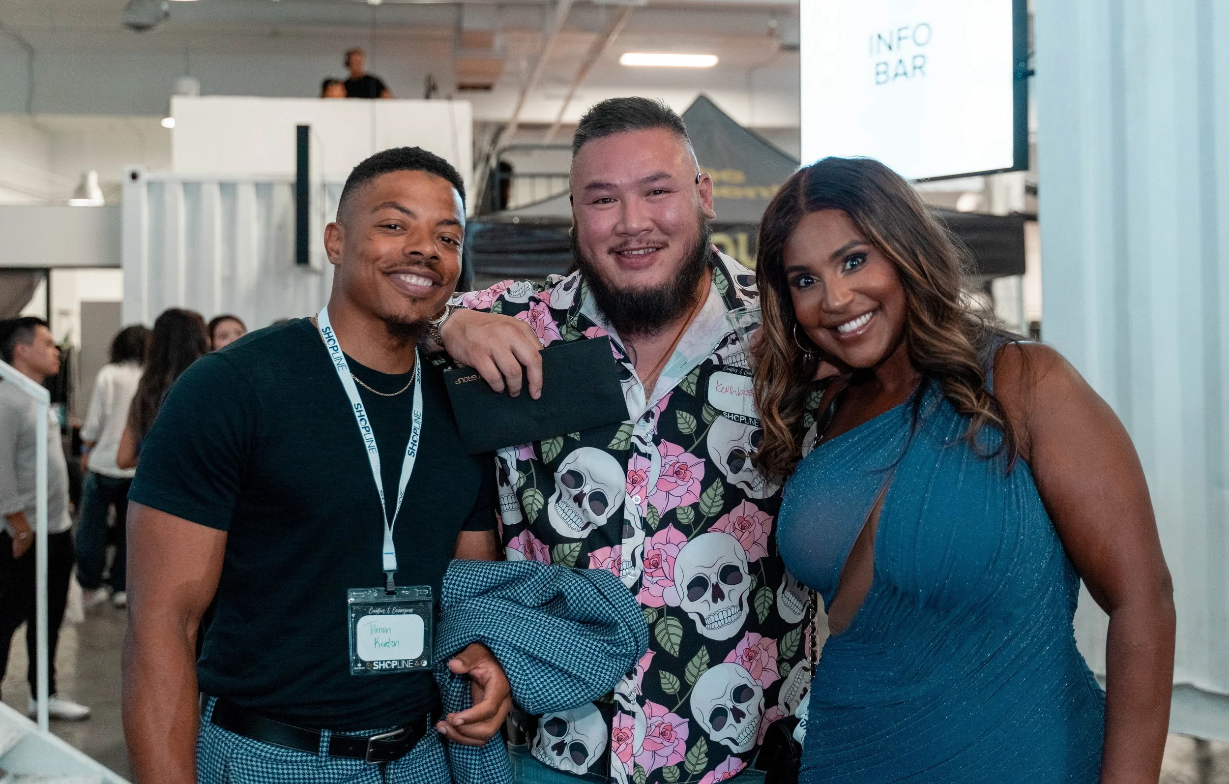 Three smiling people posing together at an event with a crowd and a display screen in the background.