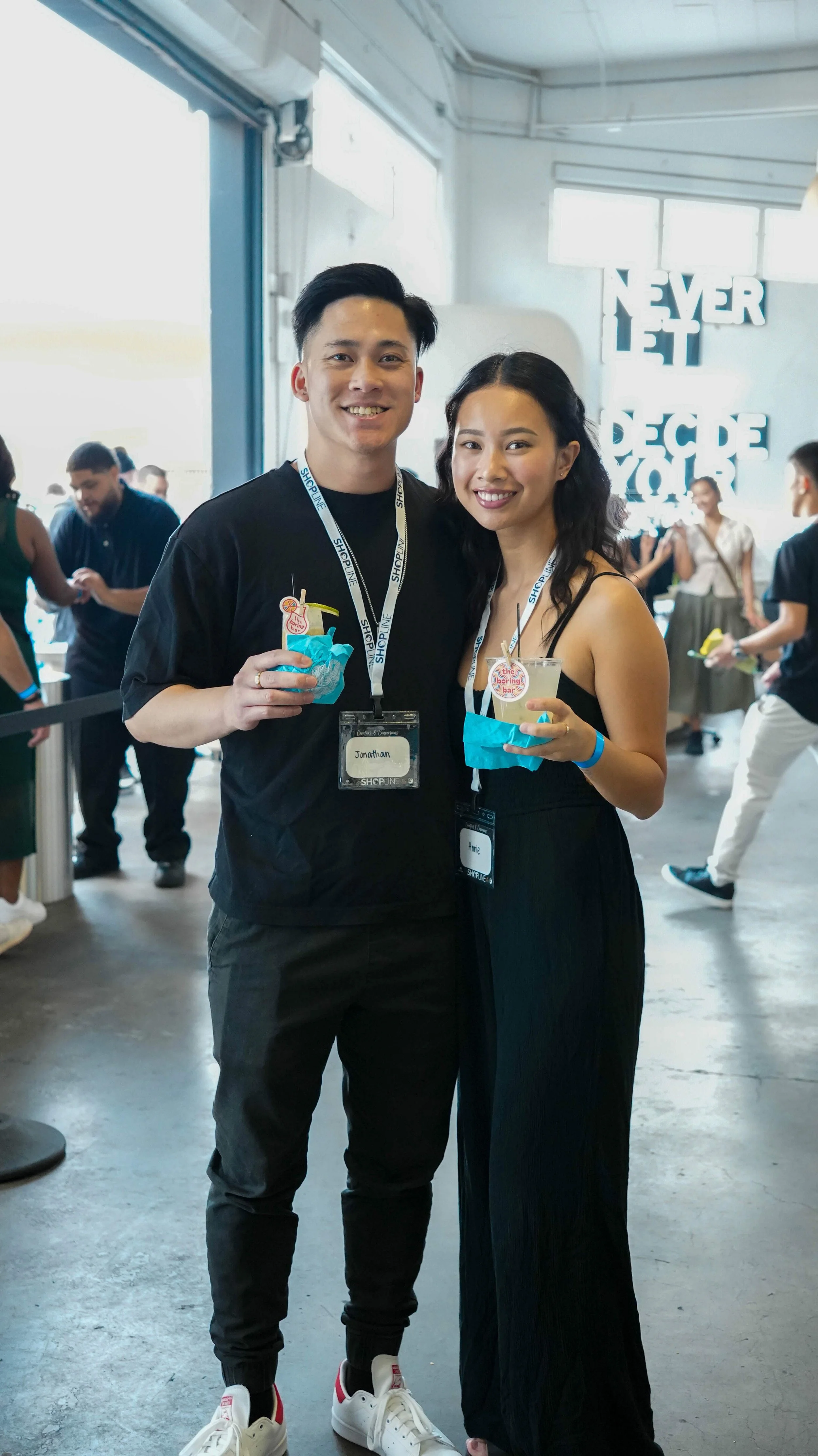 Two people, a man and a woman, smiling and holding drinks at a social event indoors, with a sign that says "Never Let Decipher" in the background.