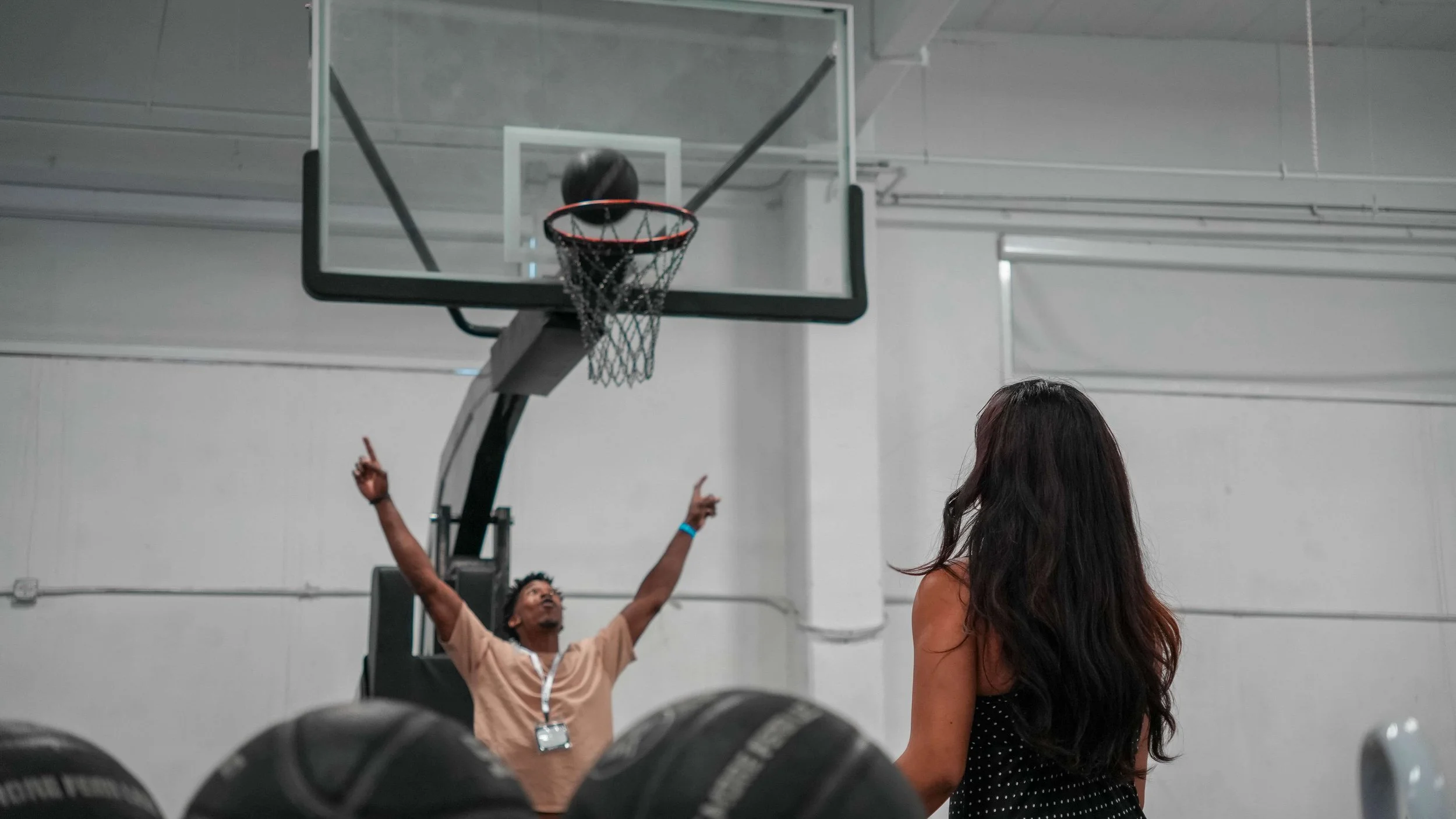 A man with glasses and a beige shirt shooting a basketball into a hoop while a woman with long dark hair watches.