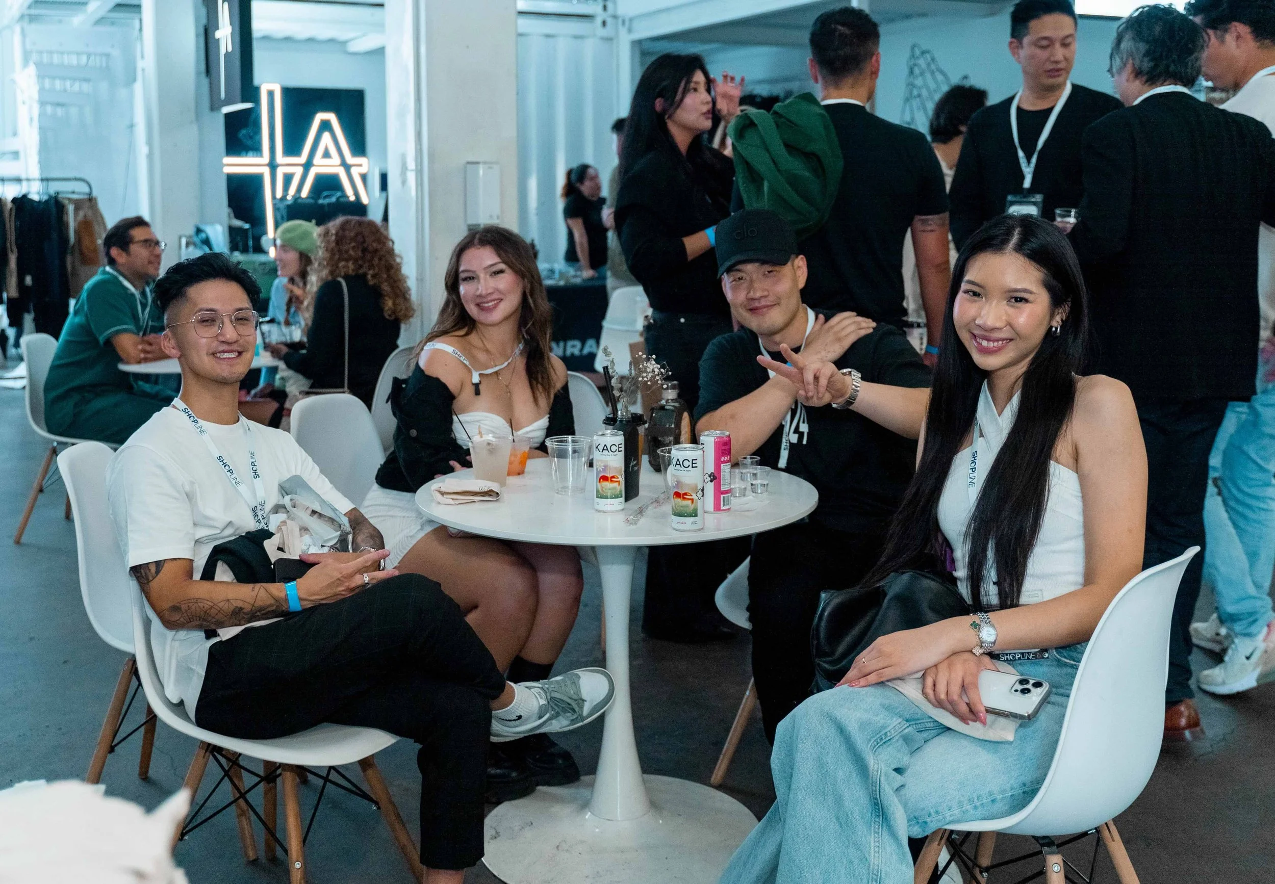 Four young adults sitting around a small round table at an indoor event, smiling at the camera. The group includes two women and two men, with various drinks and cans on the table. In the background, other people are socializing in a well-lit area wi