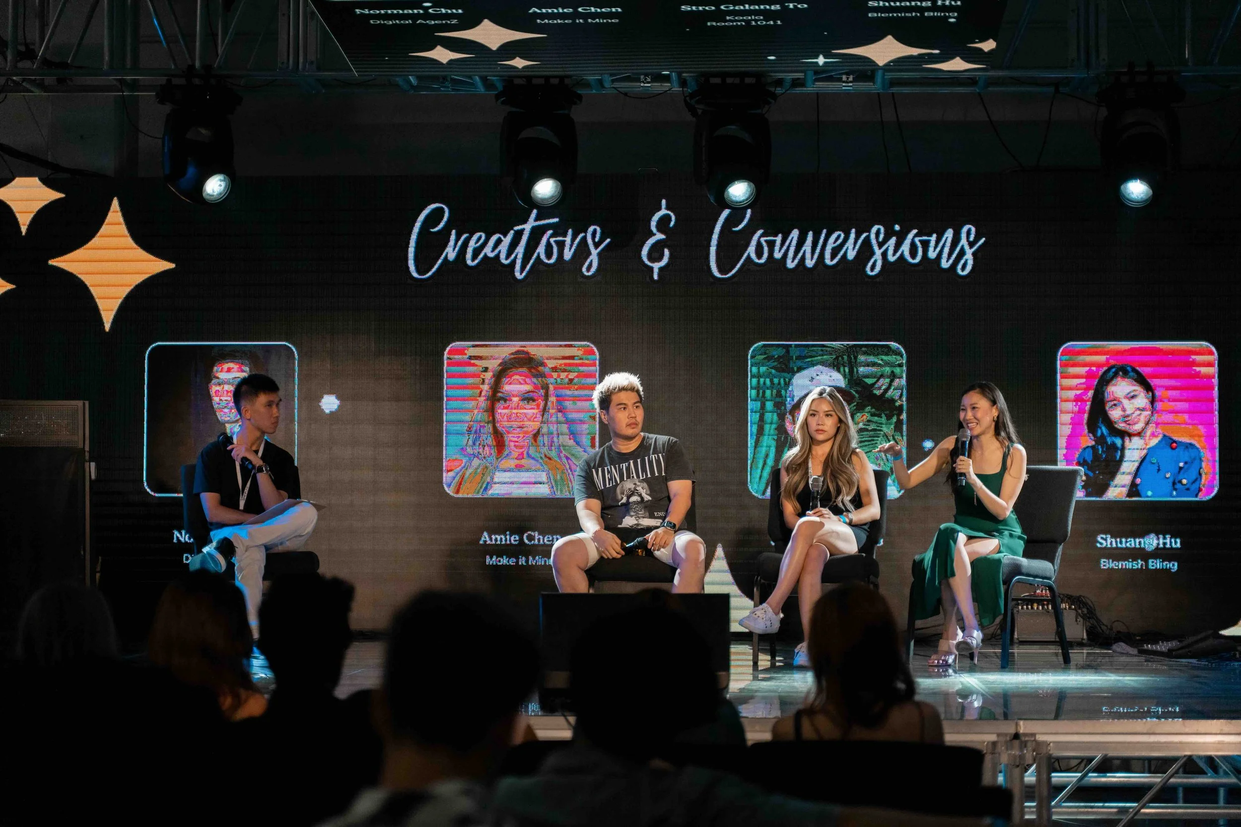 Panel discussion titled 'Creators & Conversions' with four speakers on a stage, two women and two men, in a well-lit room with an audience. The background displays colorful graphics and portraits of the speakers, with one woman speaking into a microp