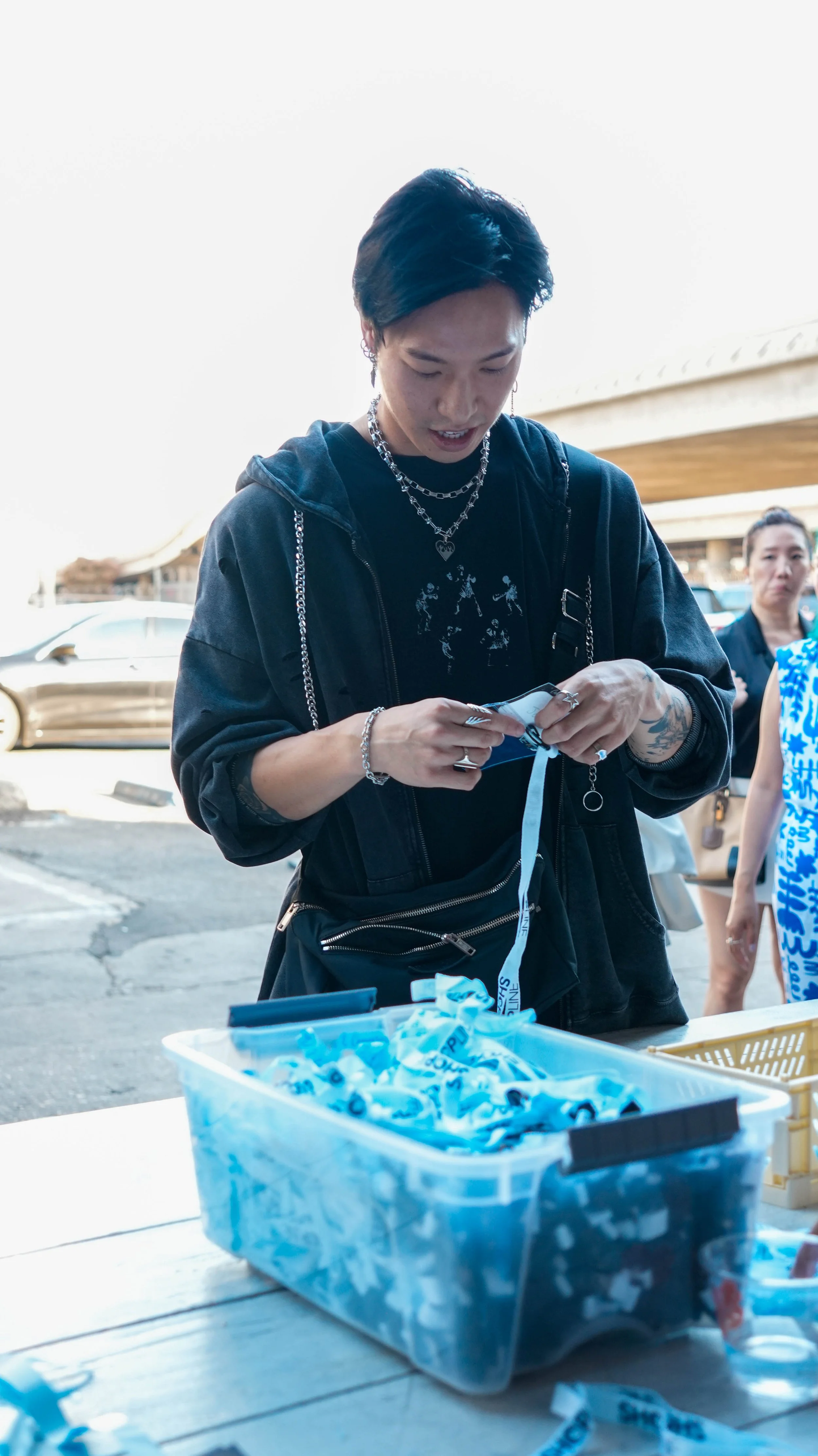 Person with short dark hair wearing black clothing and silver jewelry, working with blue wristbands at an outdoor table.