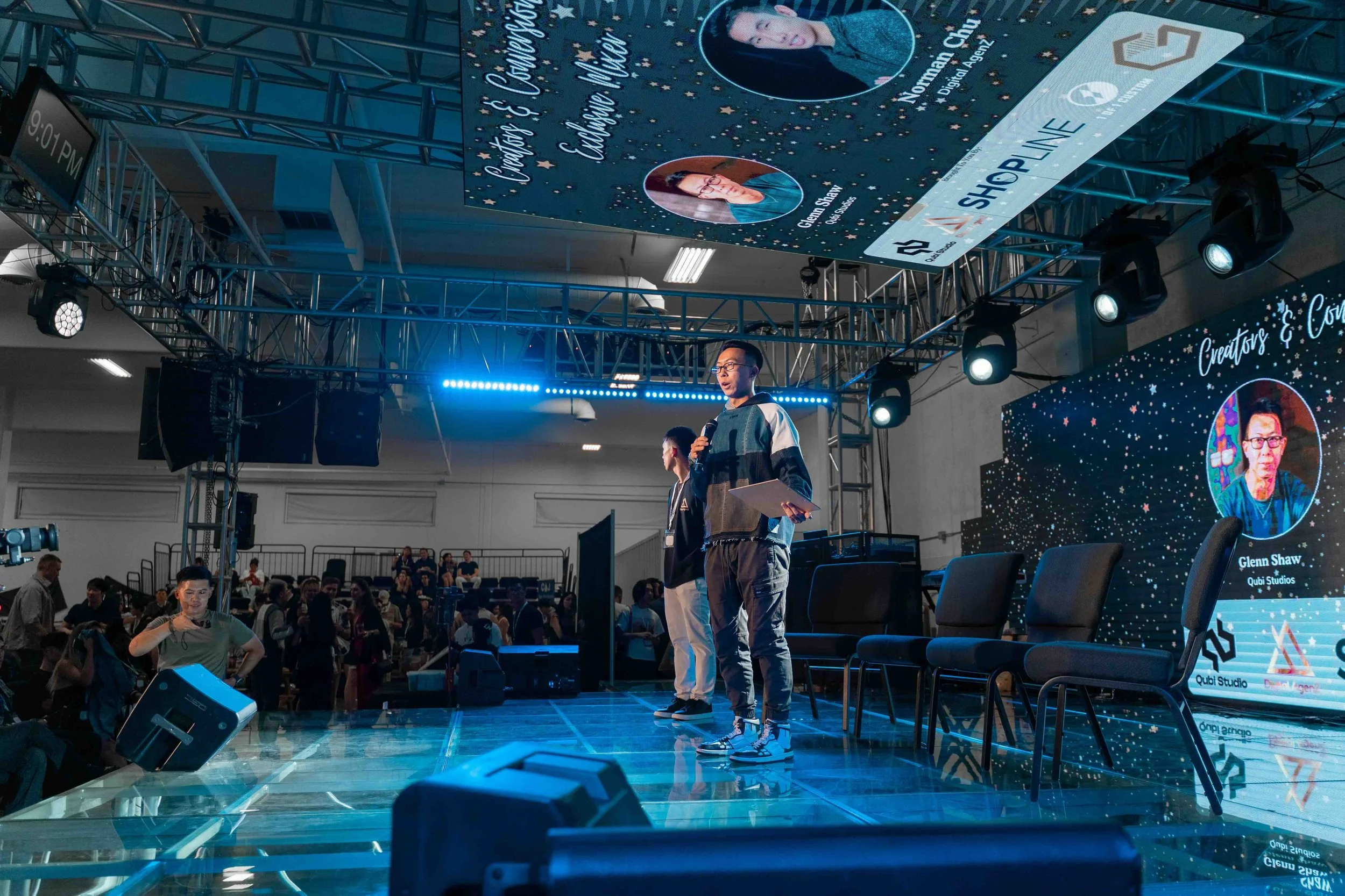Two men standing on a stage in front of an audience at a conference, with a screen displaying their names and photos behind them, surrounded by stage lights and a metal truss structure.