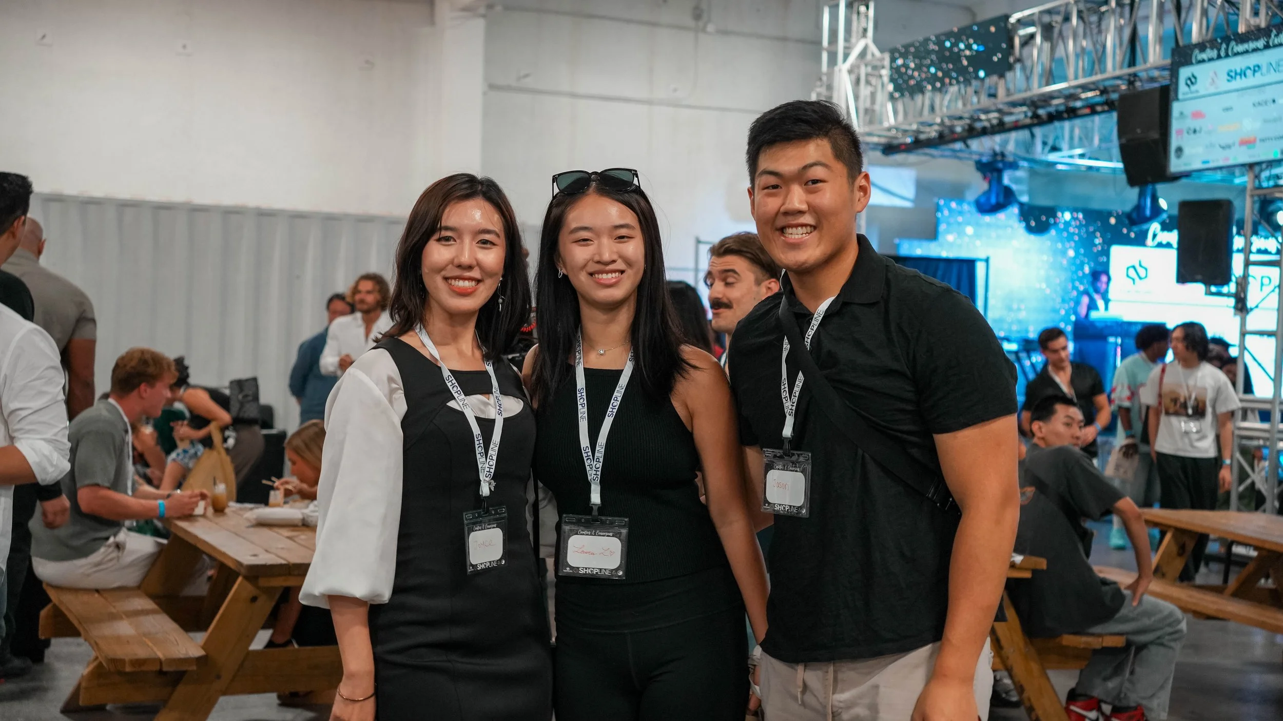 Three young adults smiling at the camera at a conference or event, wearing name tags, with a crowded background and stage setup.