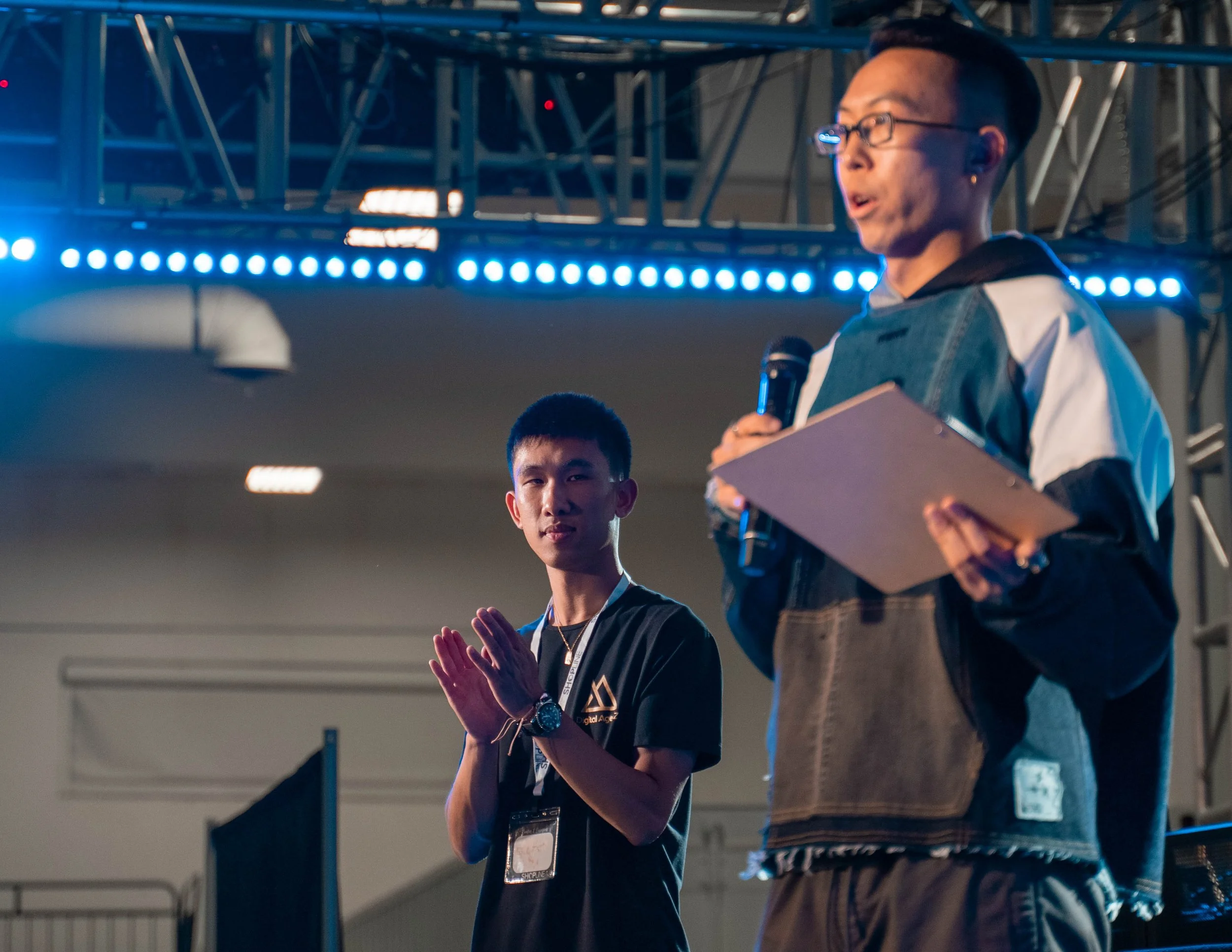 Two young men speaking at an event on a stage in a conference hall. One is holding a microphone and a clipboard, the other has hands clasped together. The stage has blue LED lighting and metallic scaffolding overhead.