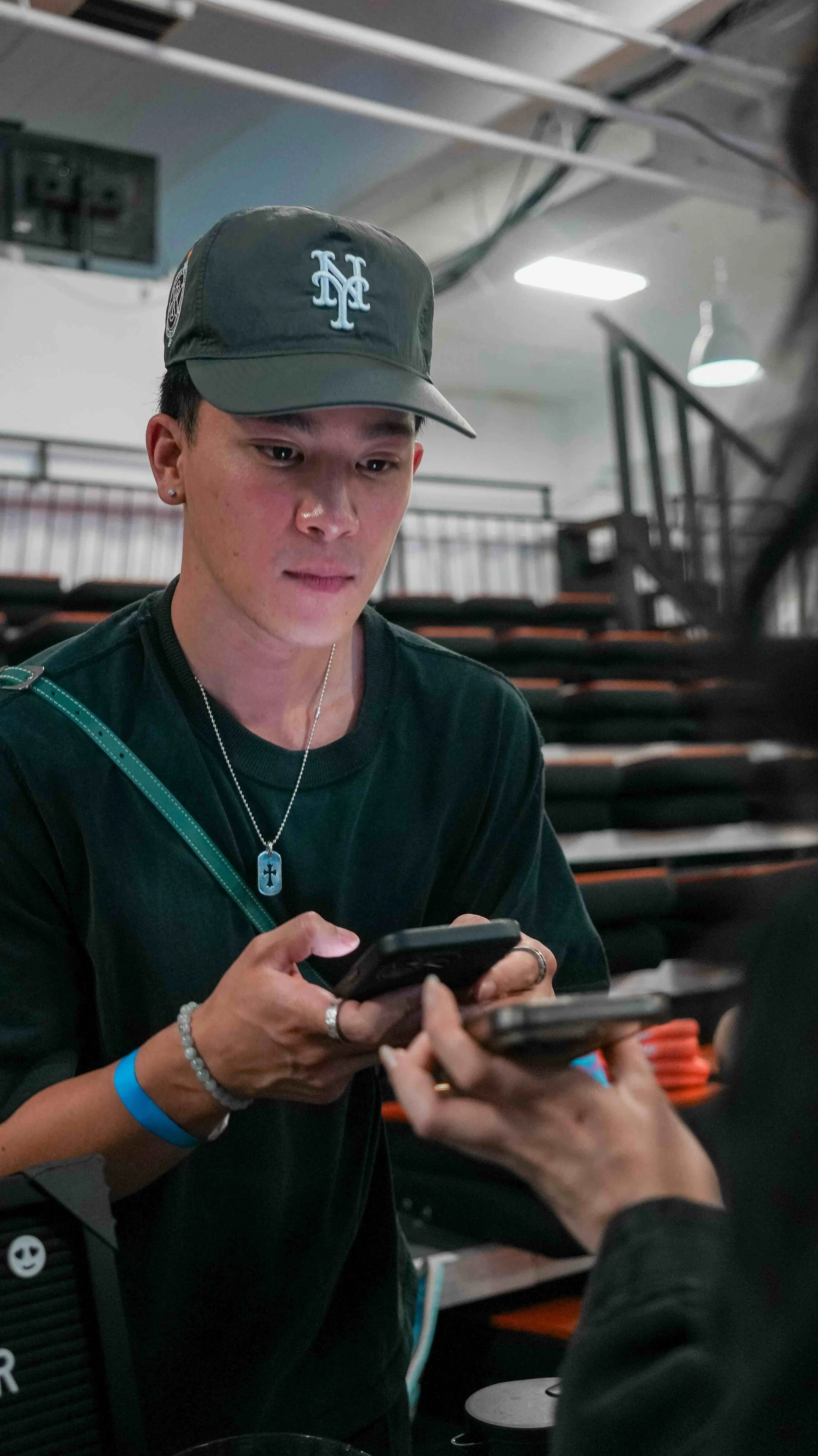 A young person wearing a baseball cap and multiple necklaces, looking at a phone, while someone else hands them a phone in a room with stacked chairs and industrial lighting.
