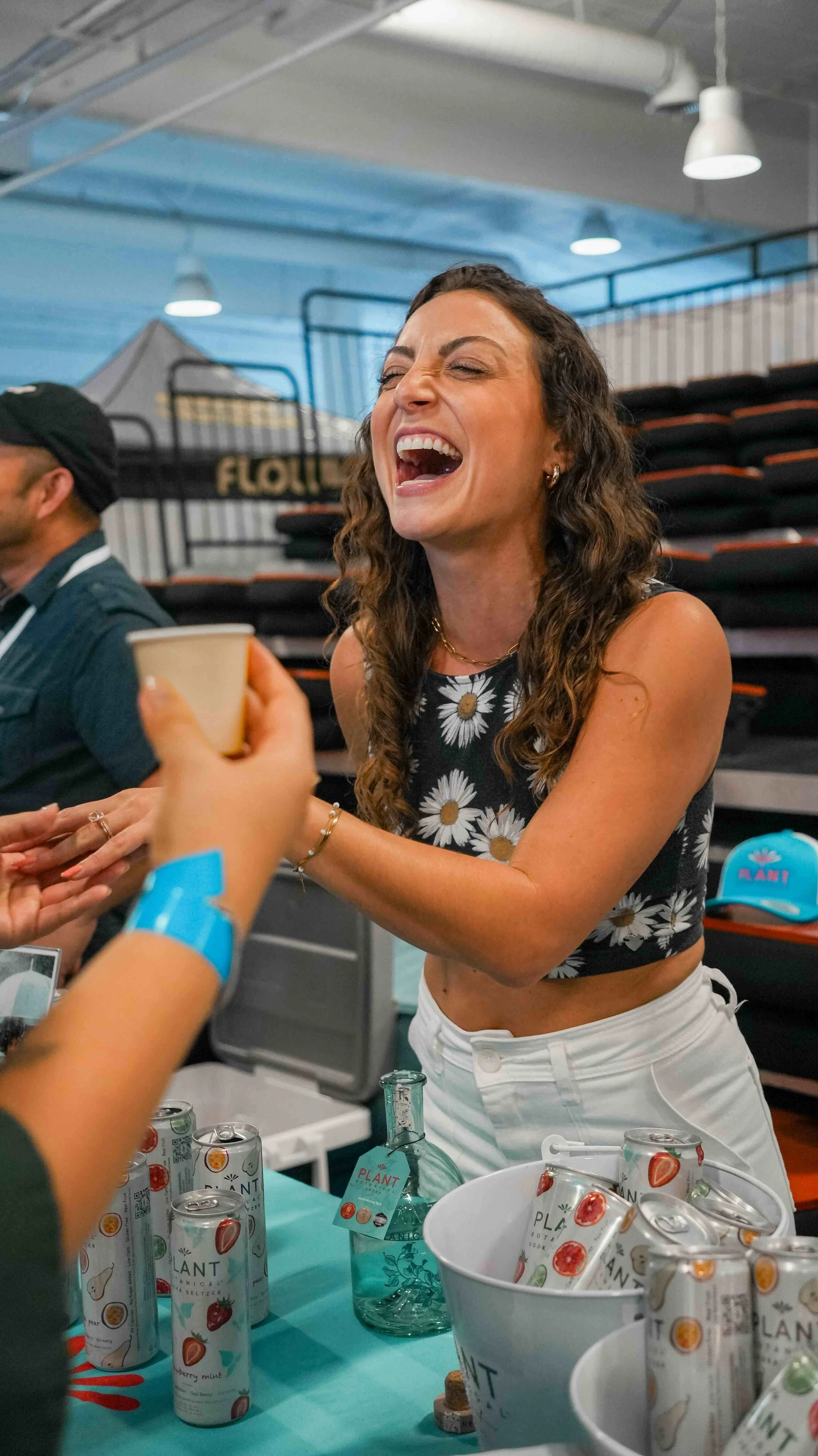A woman with long curly hair laughing and holding a cup, wearing a black crop top with white daisies and white pants, at an indoor event with cans of drink and a turquoise tablecloth.