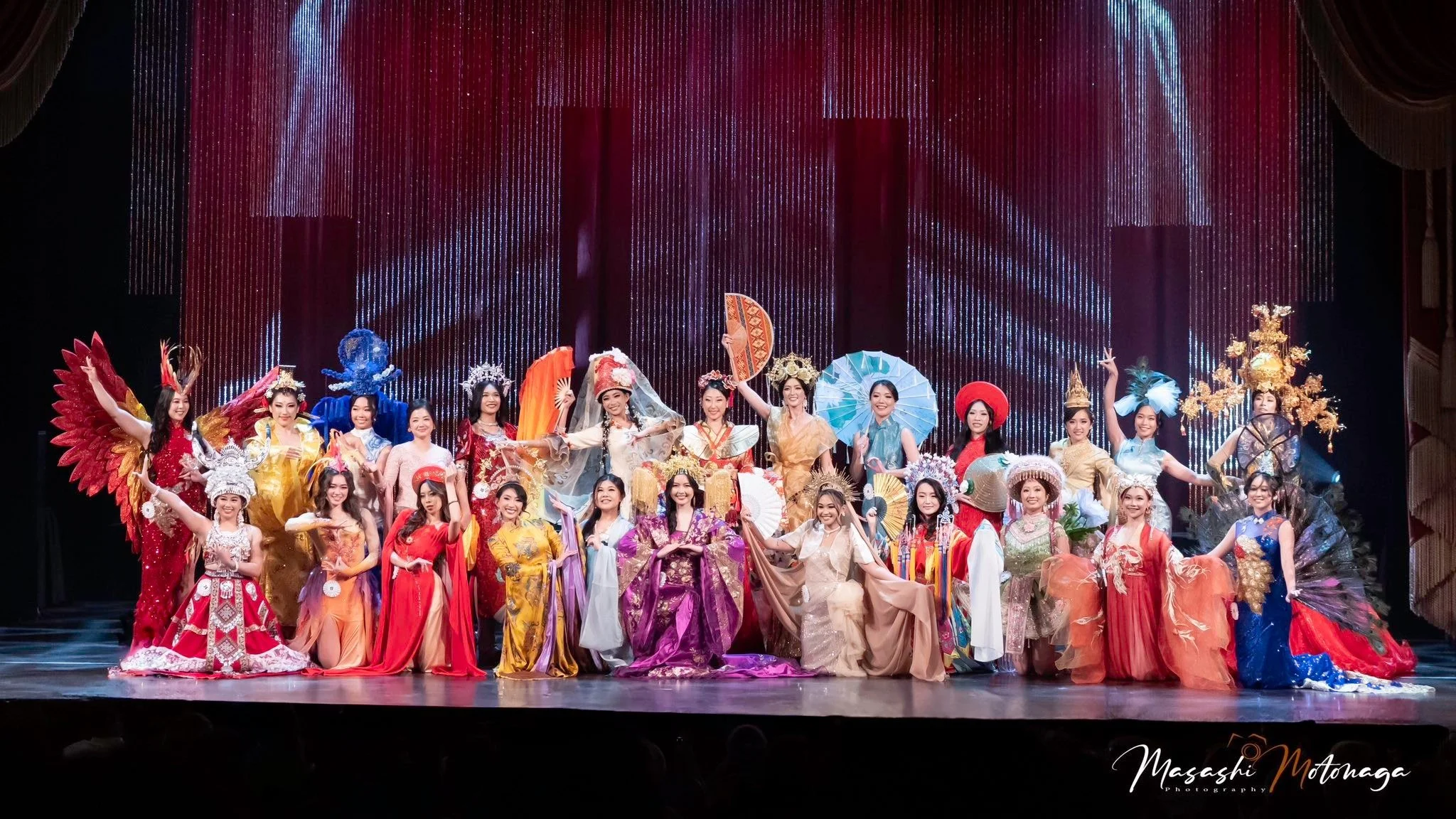 Group of women in colorful traditional costumes and accessories on stage for a cultural performance, with a decorated backdrop and stage lights.