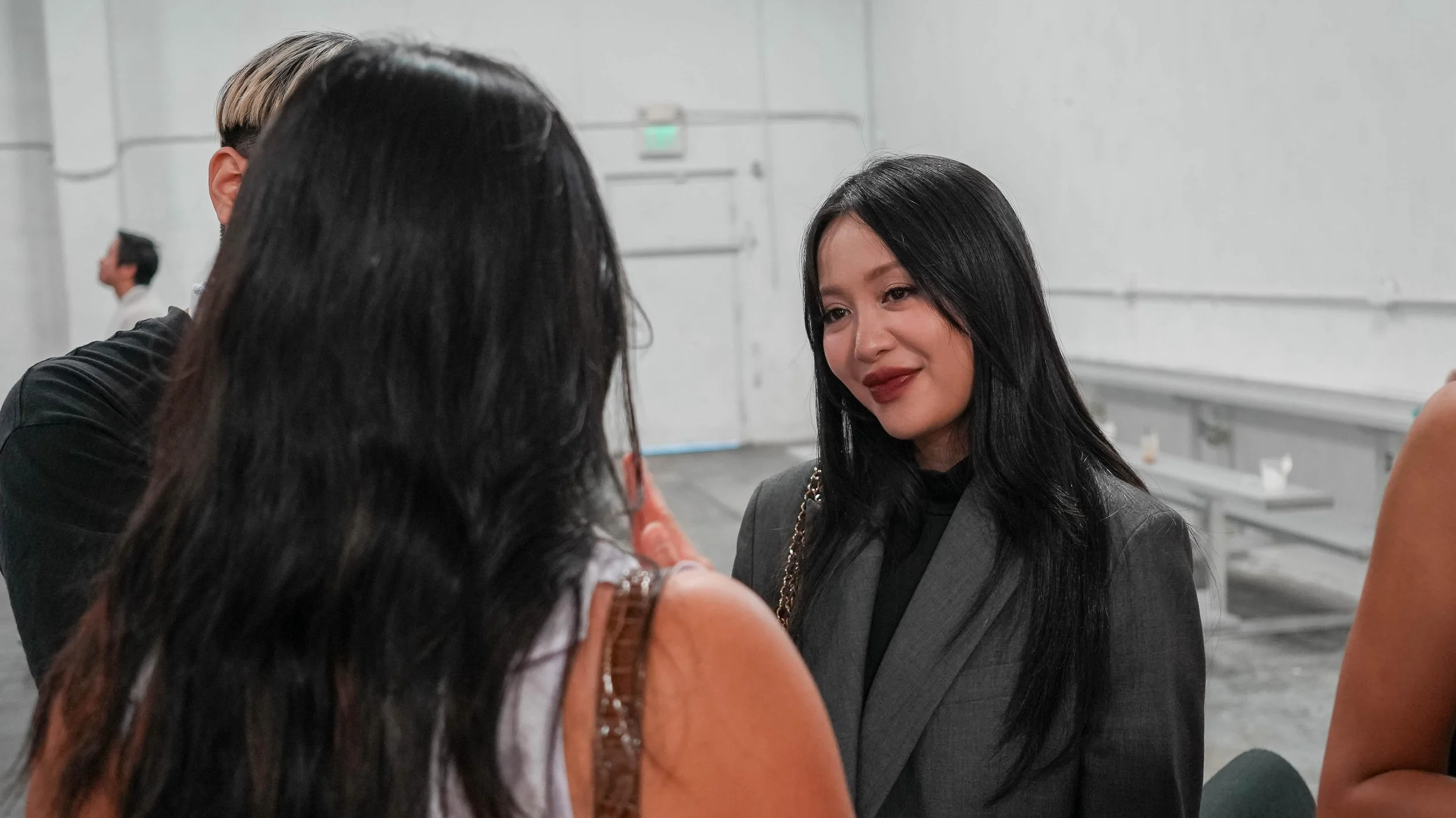 A woman with black hair and makeup smiling, wearing a gray blazer, talking to two other women with long dark hair in an indoor setting.