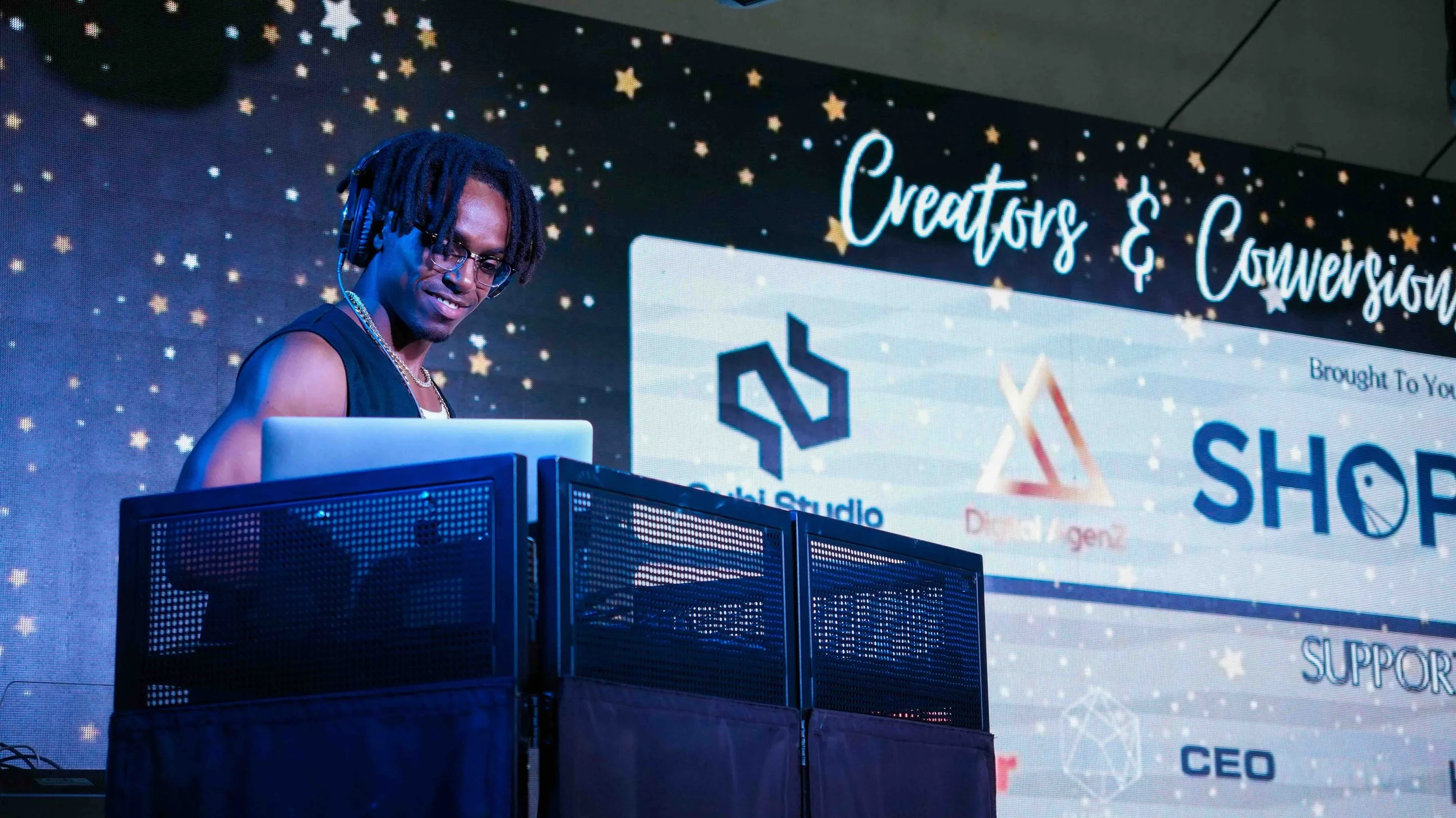 A DJ with headphones and glasses performing at an event with a large screen behind him displaying the words 'Creators & Convergence' and logos of sponsors.