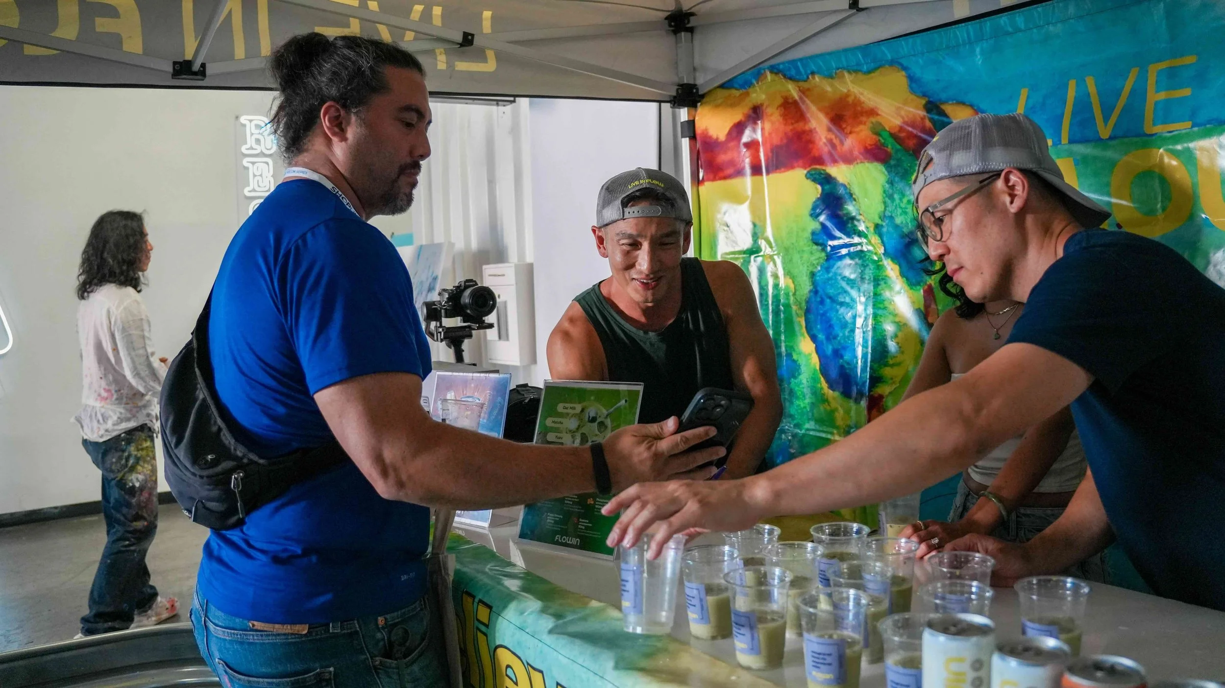Three men at an outdoor booth looking at a smartphone and various samples of greenish liquids in small cups. A colorful rainbow background is visible behind them.
