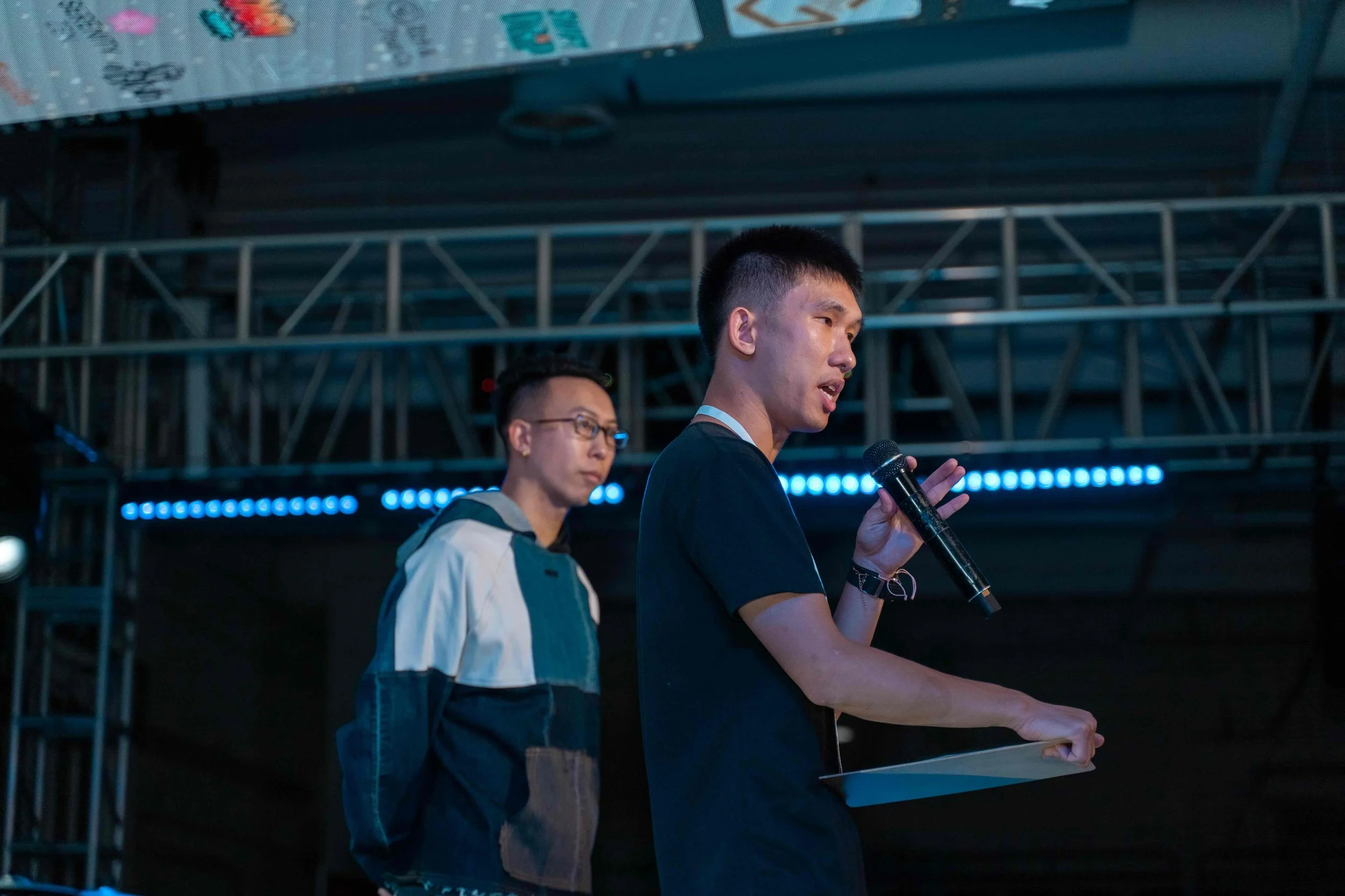 Two young men on stage, one speaking into a microphone while holding a clipboard, the other standing behind him, with stage lighting and metal scaffolding in the background.