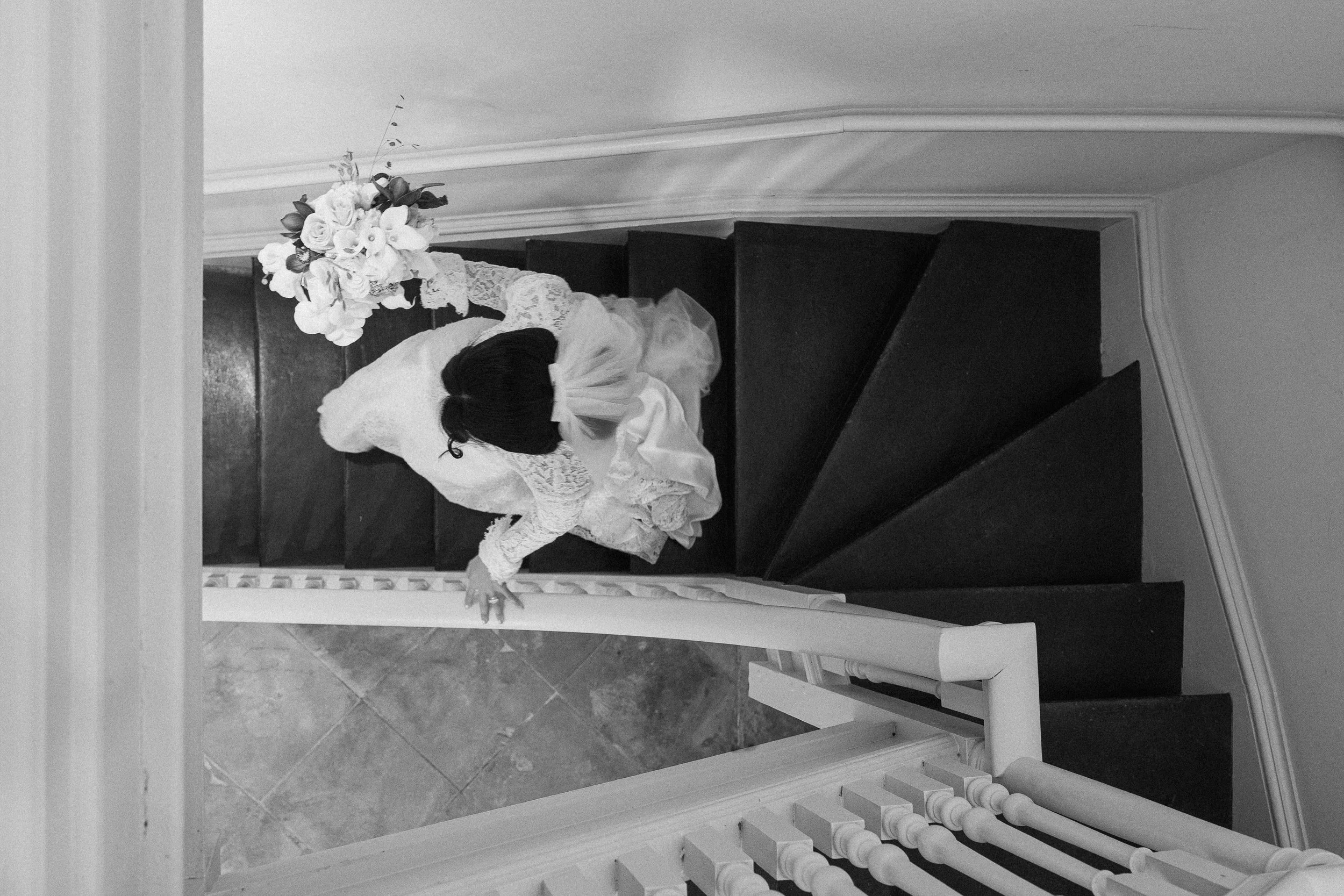 A couple, dressed in wedding attire, holding hands and walking across a grassy hill at sunset, with mountains in the background.