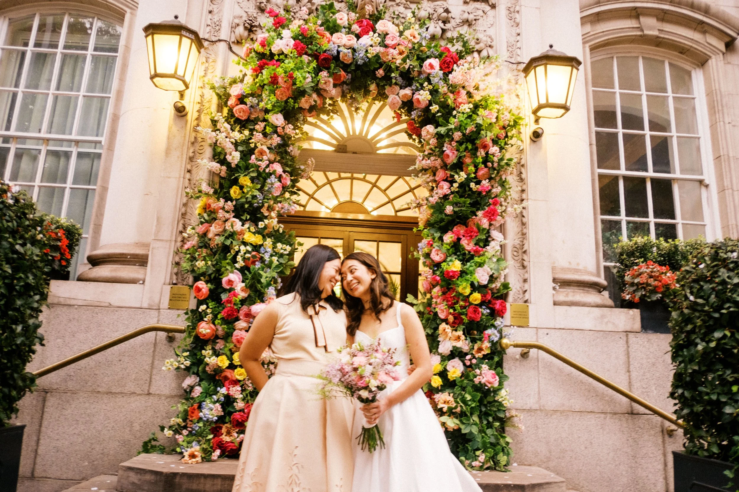 A couple holding a bouquet of orange flowers and greenery while standing outdoors in a grassy area with sunlight shining behind them.