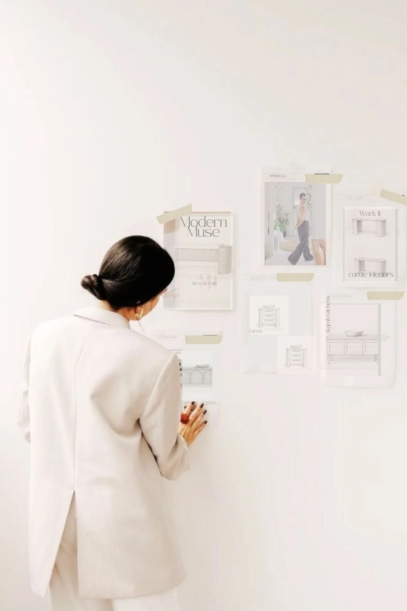 Woman in white suit looking at a wall with interior design sketches and photos taped with beige tape.