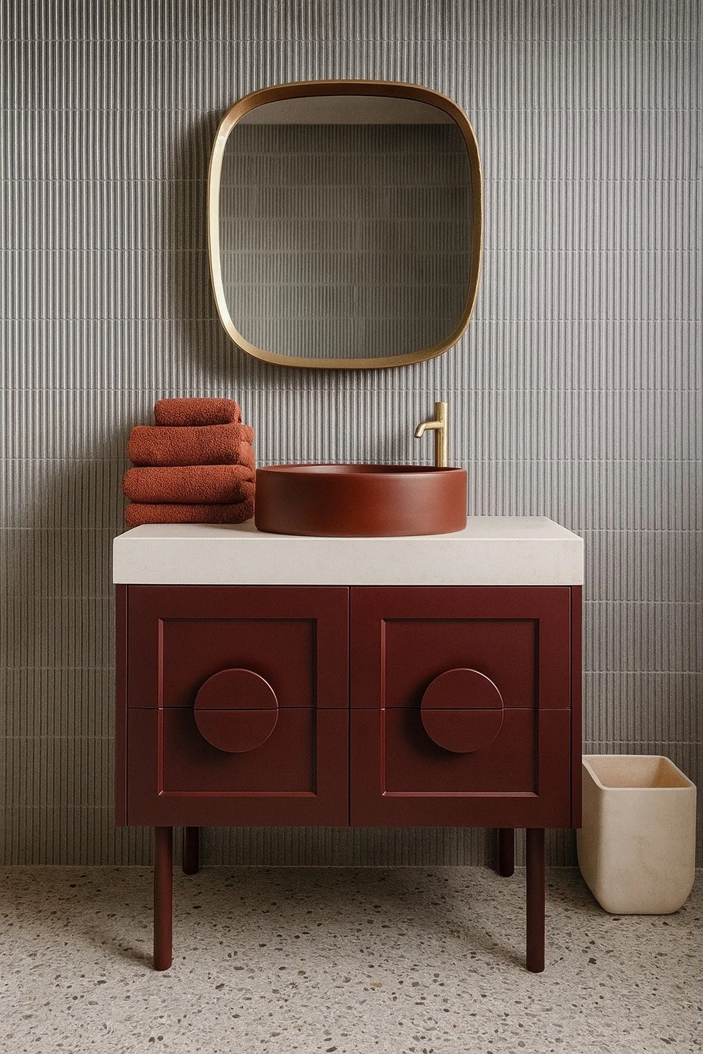 A modern bathroom vanity with a red cabinet, white countertop, and round red sink. Above it is a large, rounded rectangle mirror with a gold frame. To the left of the sink are three folded red towels, and to the right is a beige container on the terrazzo floor. The wall behind has gray textured tiles.