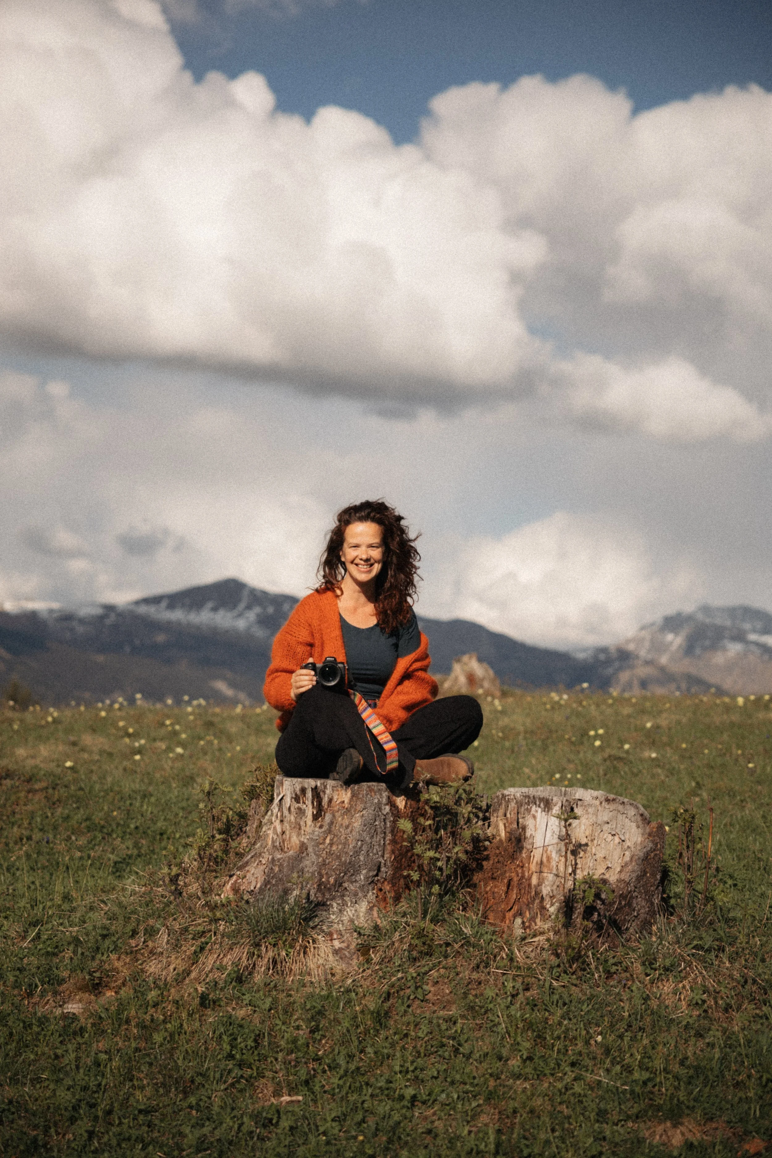 Eine lachende Frau sitzt auf einem Baumstumpf in einer grünen Wiese mit Bergen im Hintergrund, hält eine Kamera in der Hand und trägt einen orangefarbenen Pullover.