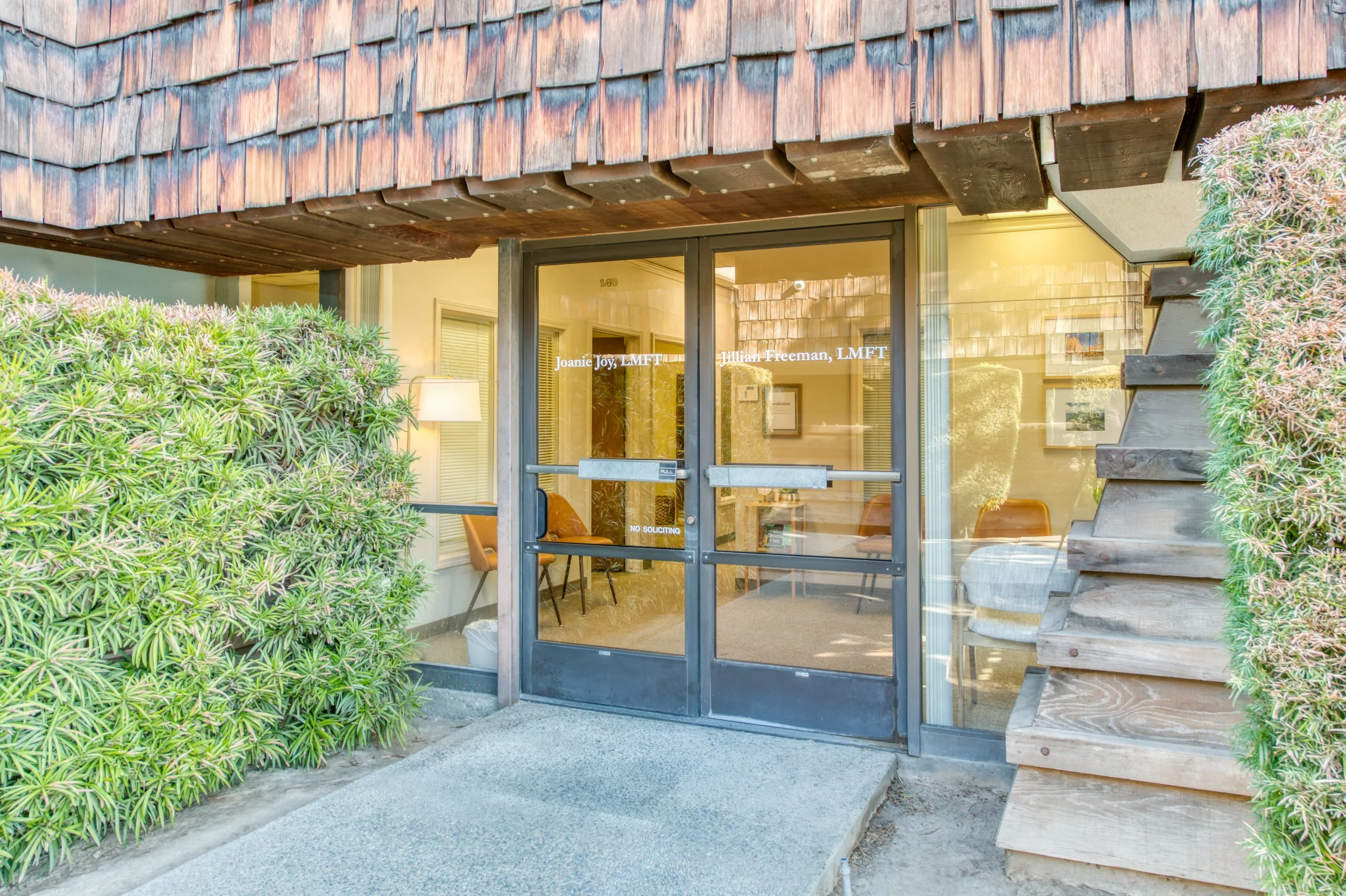 The entrance to a professional office with glass double doors, small bushes on either side, and a wooden siding top, with visible interior chairs and a lamp.