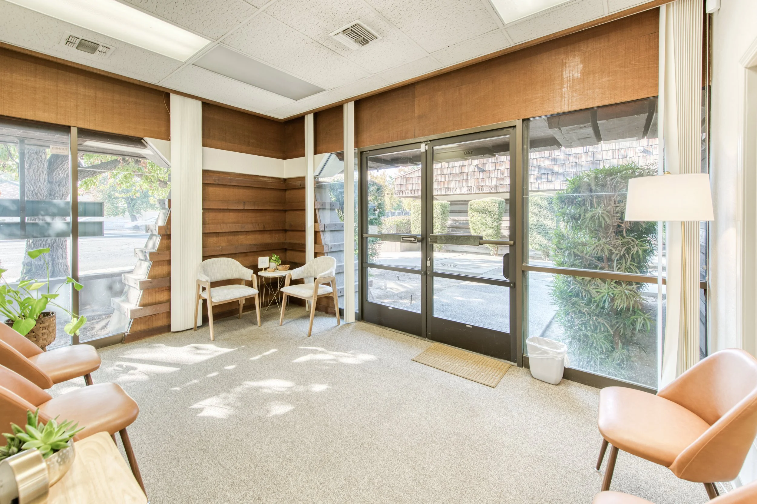 Interior view of a waiting area with chairs, a lamp, and large glass doors and windows showing outdoor greenery.