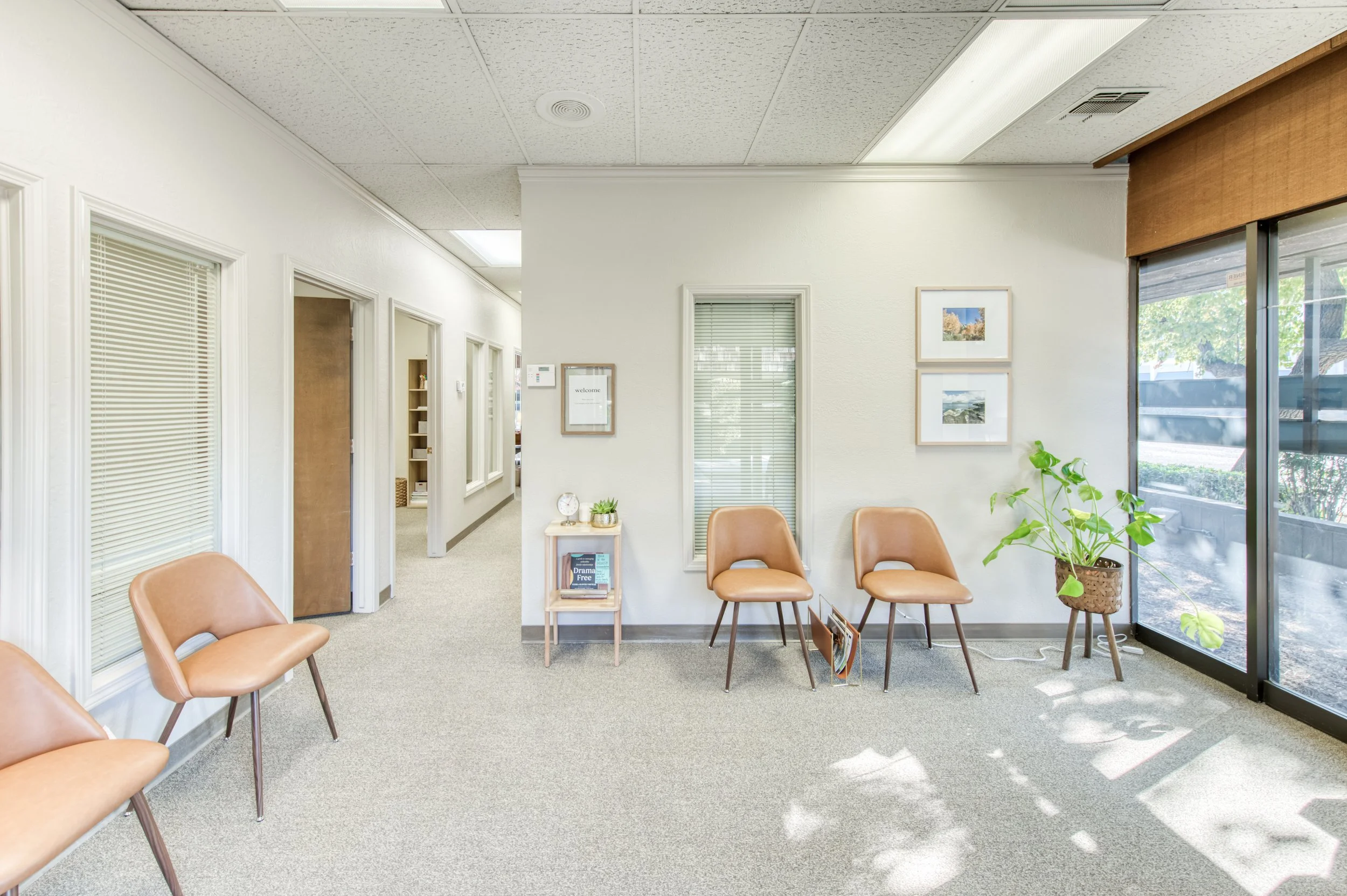 Waiting room with four tan chairs, a potted plant, and framed pictures on the wall, near large window with sunlight and blinds.