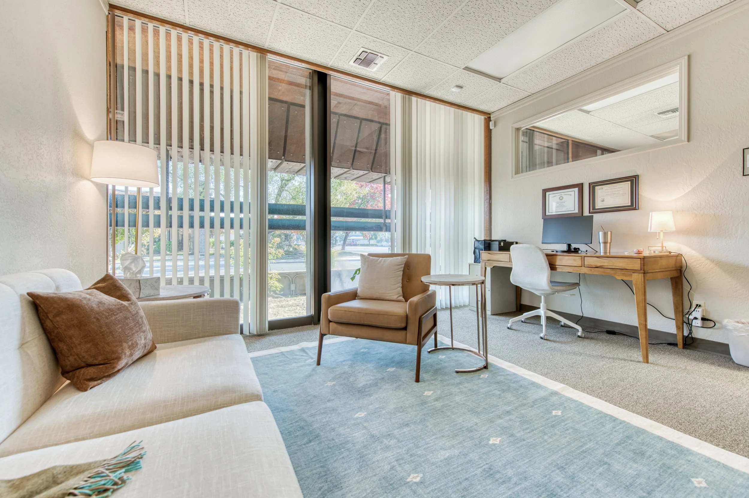 Interior of an office or waiting area with a cream sofa, brown pillows, a beige armchair, a wooden desk with a computer, a white chair, and large windows with vertical blinds.