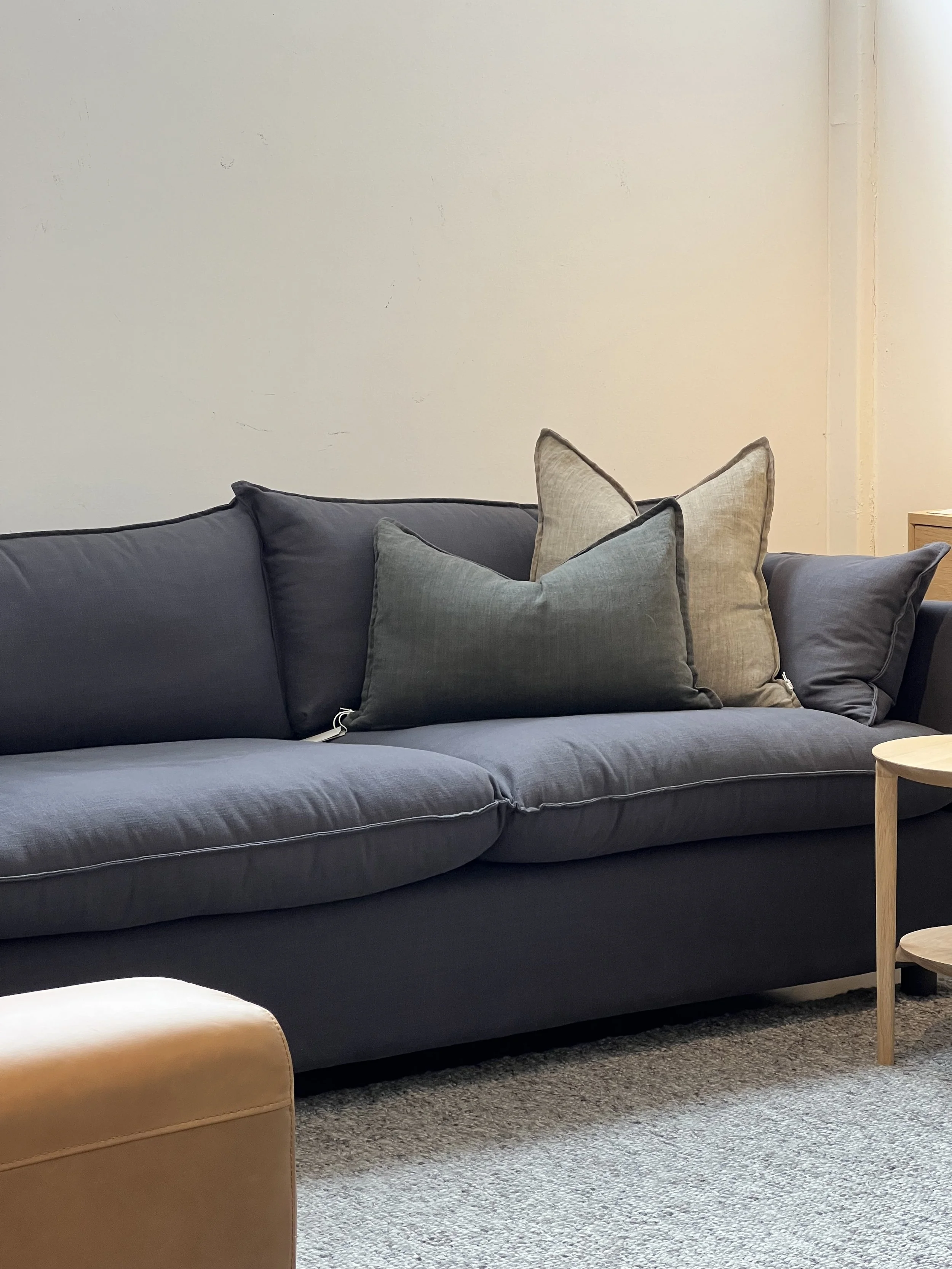 A dark grey sofa with three decorative throw pillows in varying shades of grey and beige, situated in a room with a light grey wall, a beige ottoman in the foreground, and a round wooden side table partially visible on the right.