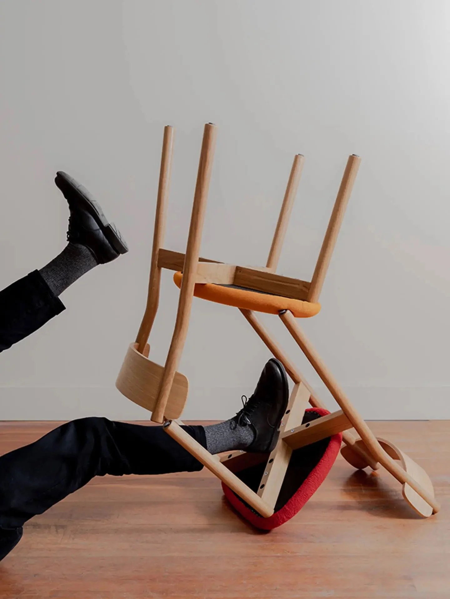 A person falls backward over a tipped over wooden chair, with their legs and feet in the air and the chair collapsing on the floor.
