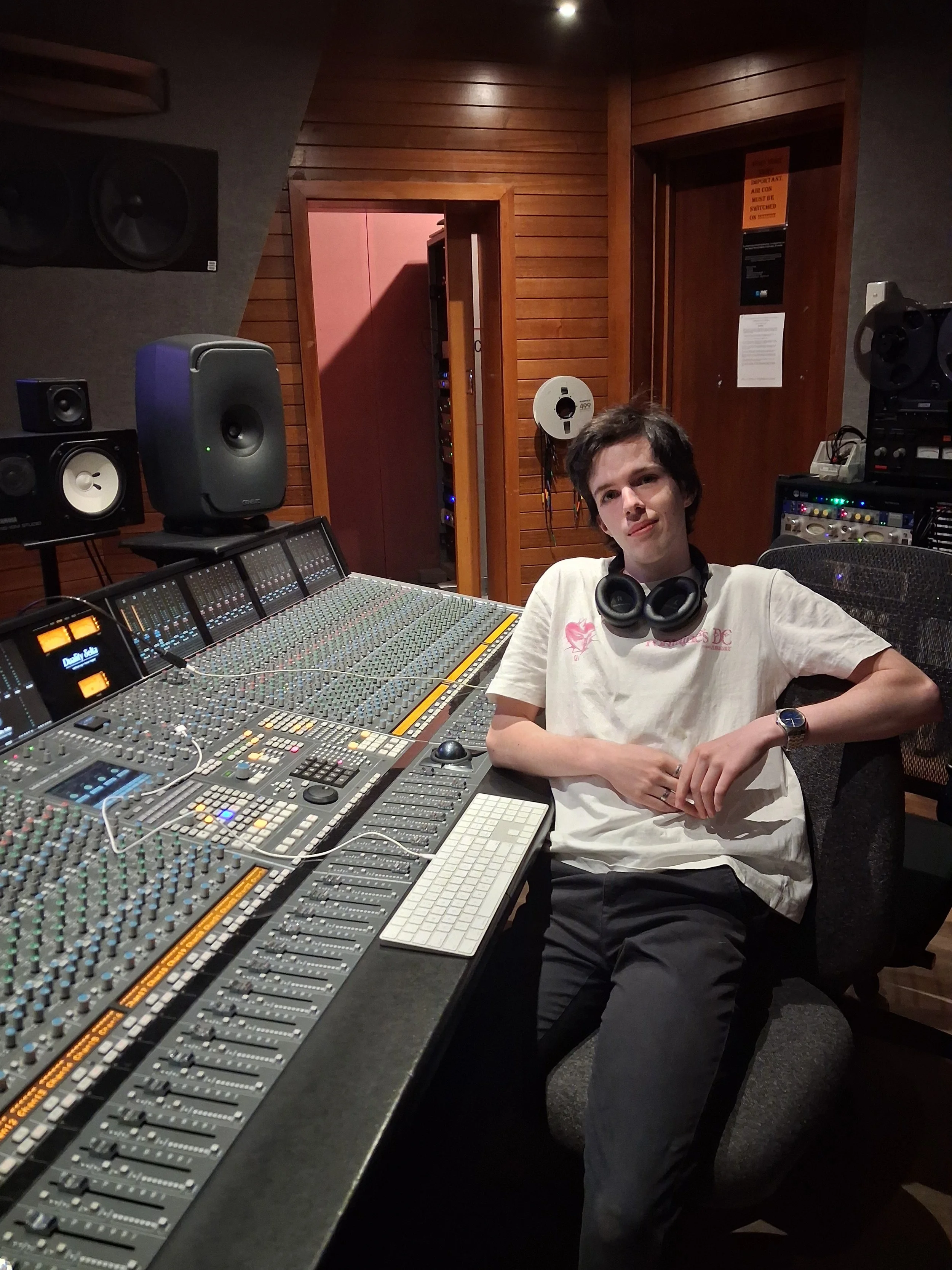 A young man with dark hair sitting in a recording studio at a large mixing console with studio monitors and audio equipment around him.