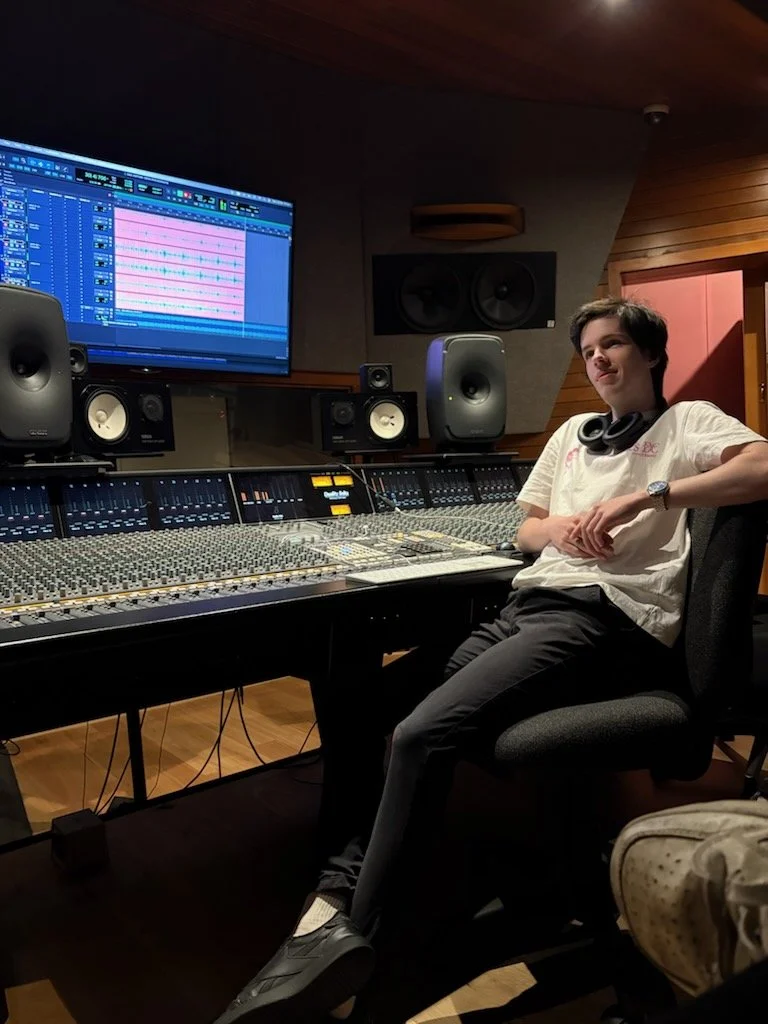 Young man sitting in a recording studio with a large mixing console, studio monitors, headphones around his neck, and a computer screen displaying digital audio workstation software.