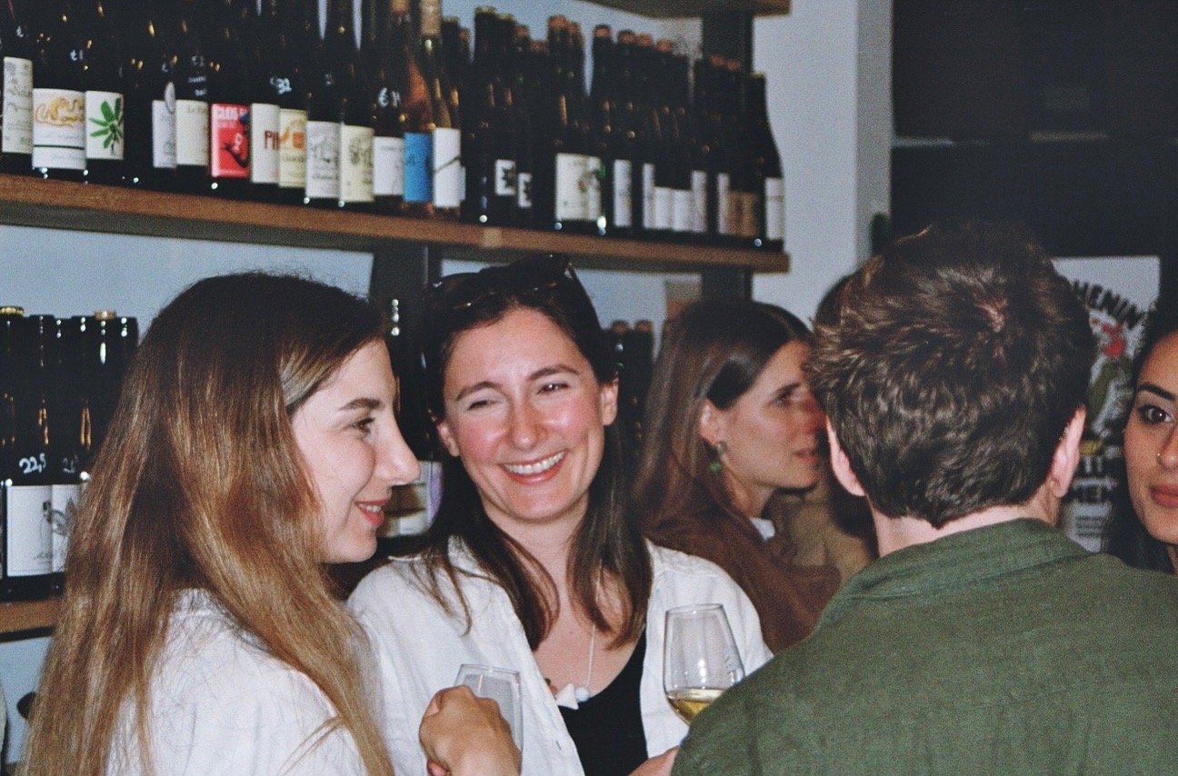 Group of people socializing at a bar, with shelves of wine bottles in the background, holding glasses of wine.