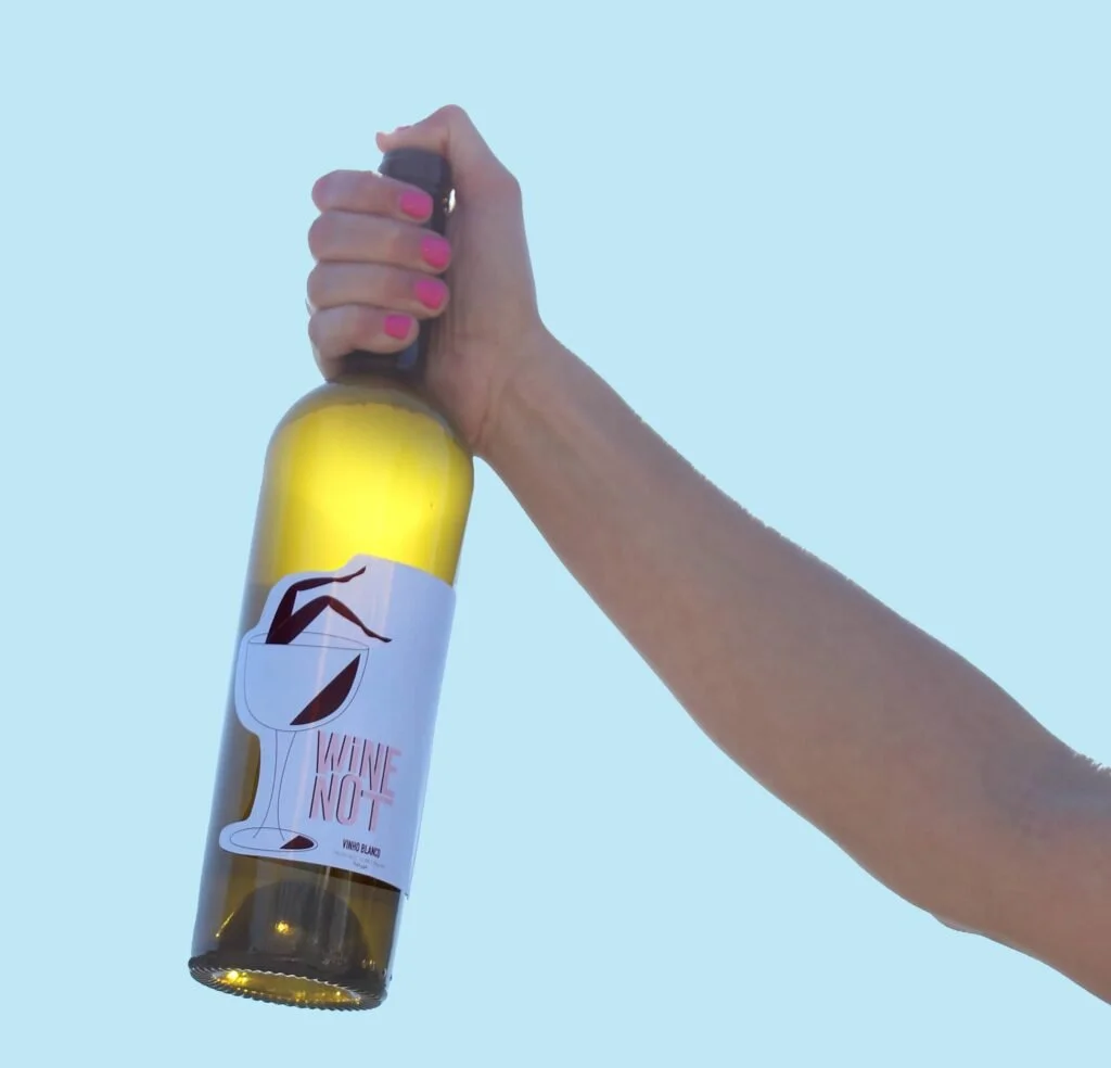 A hand with pink nail polish holding a bottle of wine with a label that says "Wine Not" and features an illustration of a wine glass with a red drink and a leaning bottle.