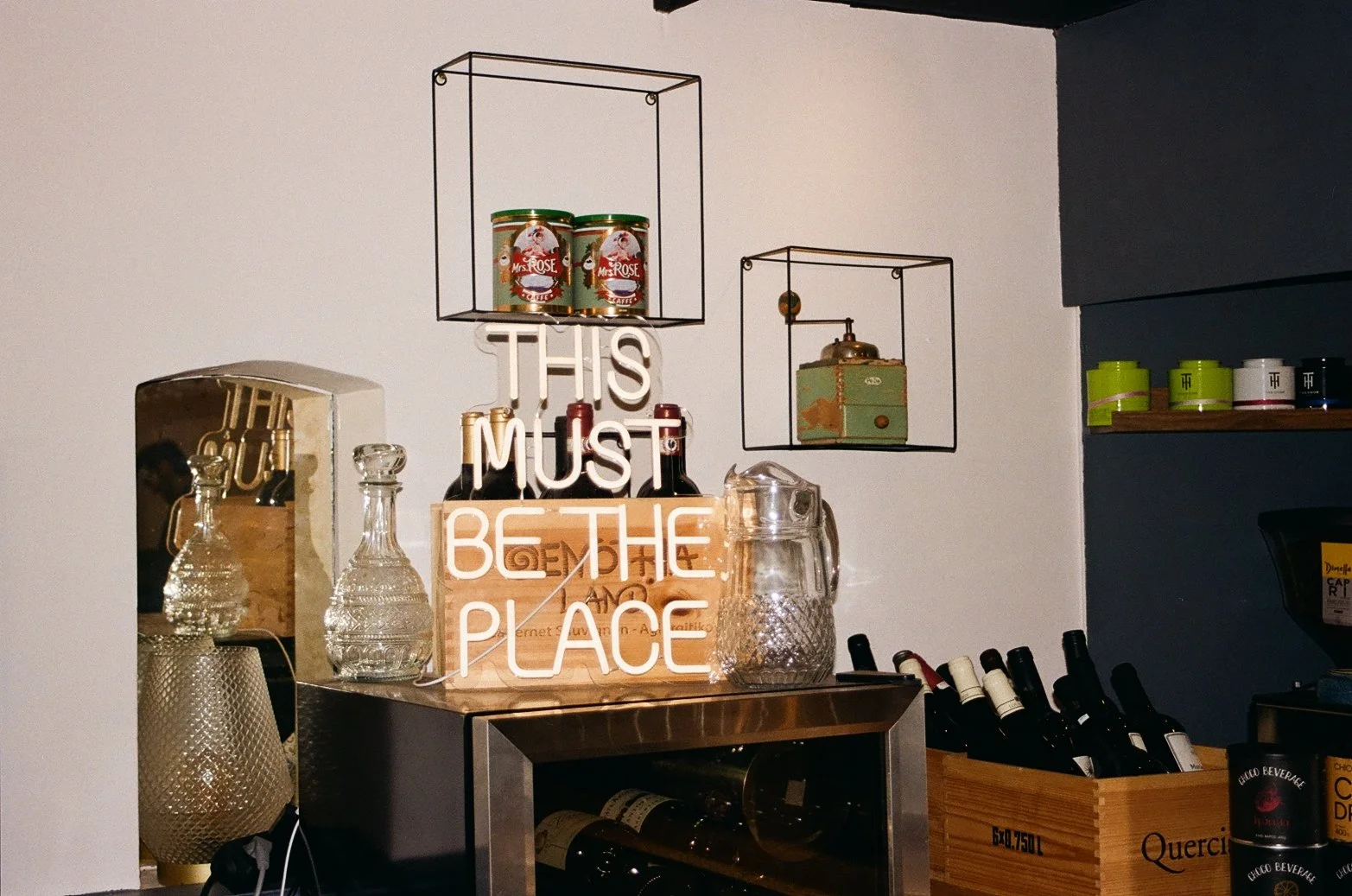 Decorative wall display with neon sign reading 'This must be the place' surrounded by glass bottles, shelves with containers, and wine bottles.