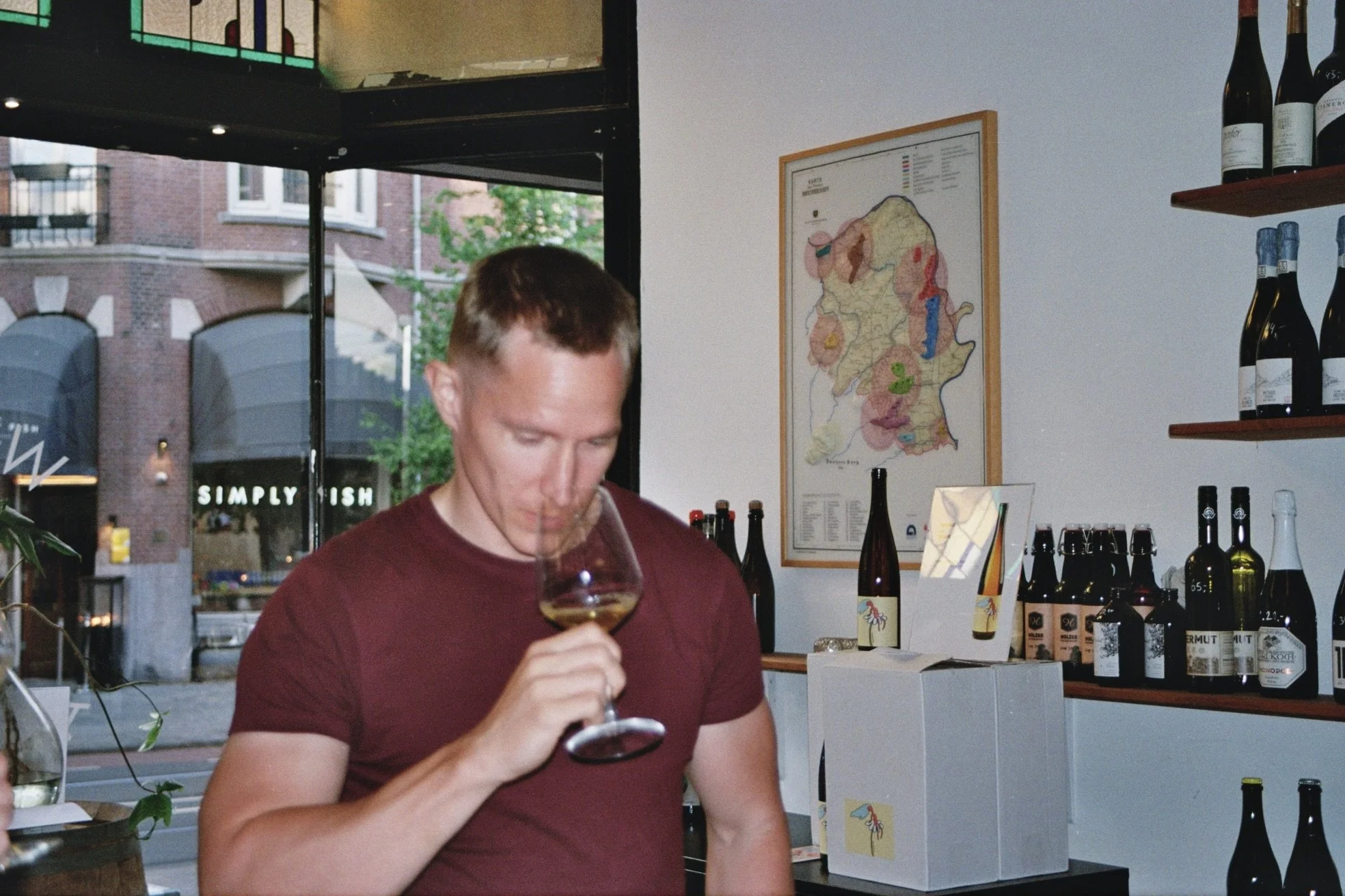 A man tasting wine in a wine shop, holding a glass of wine close to his nose, with shelves of wine bottles and a map on the wall behind him.