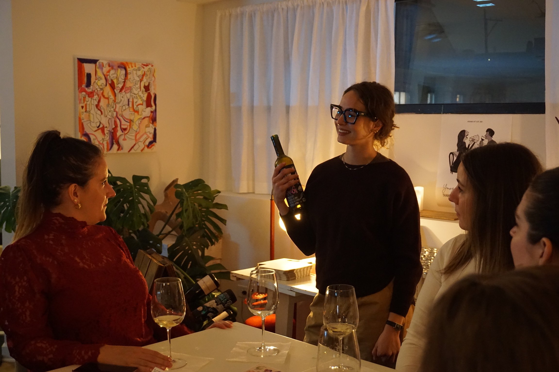 A woman in a black shirt and glasses holding a wine bottle, smiling, as she speaks to three women seated at a table, in a cozy, well-lit room with artwork on the wall.