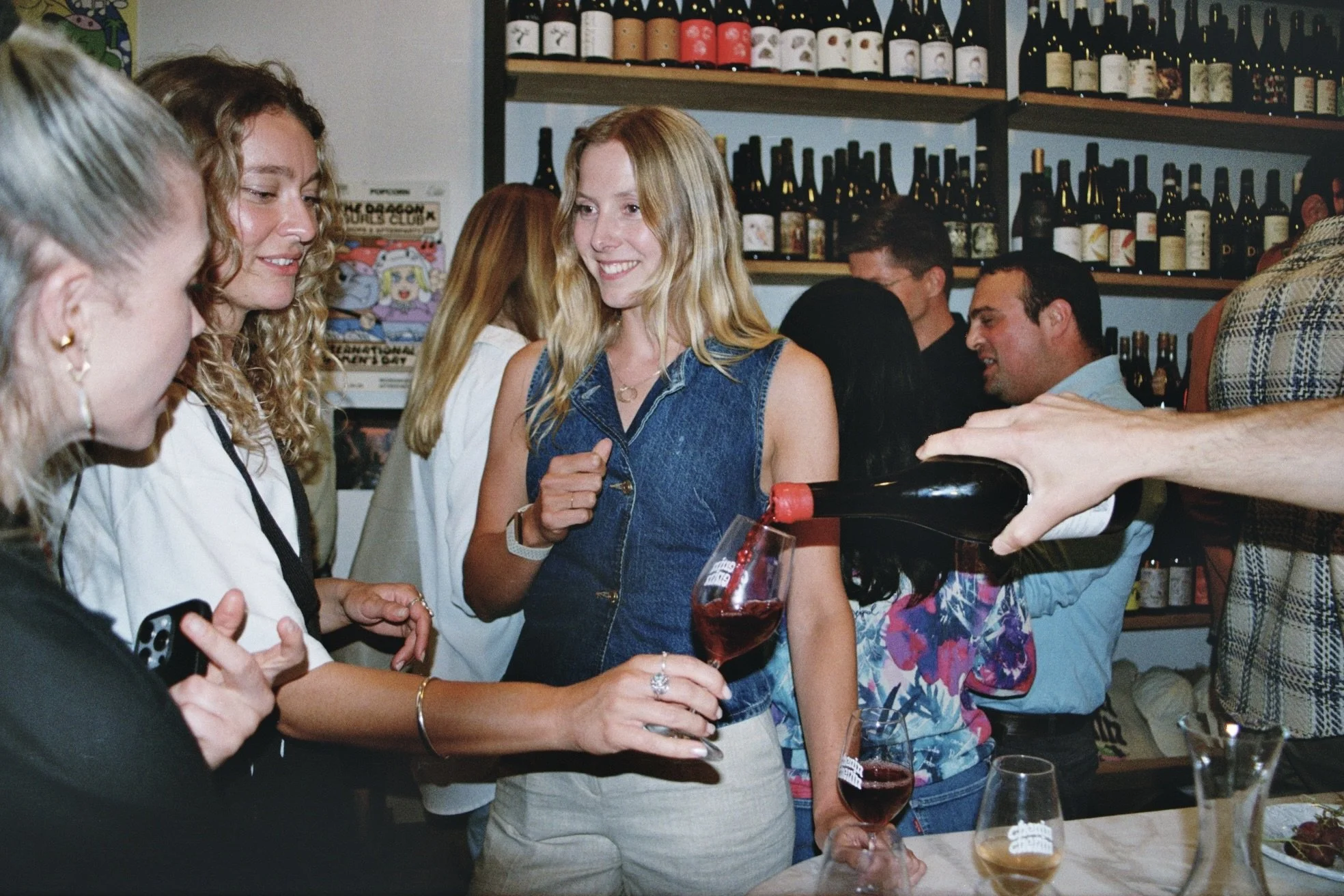 People socializing at a bar or restaurant, with a bartender pouring red wine into a glass, against shelves of wine bottles in the background.