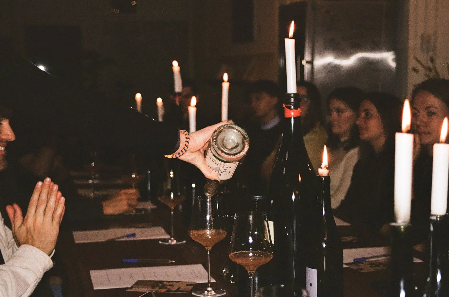 People seated at a table with lit candles, while someone pours a drink into a glass.