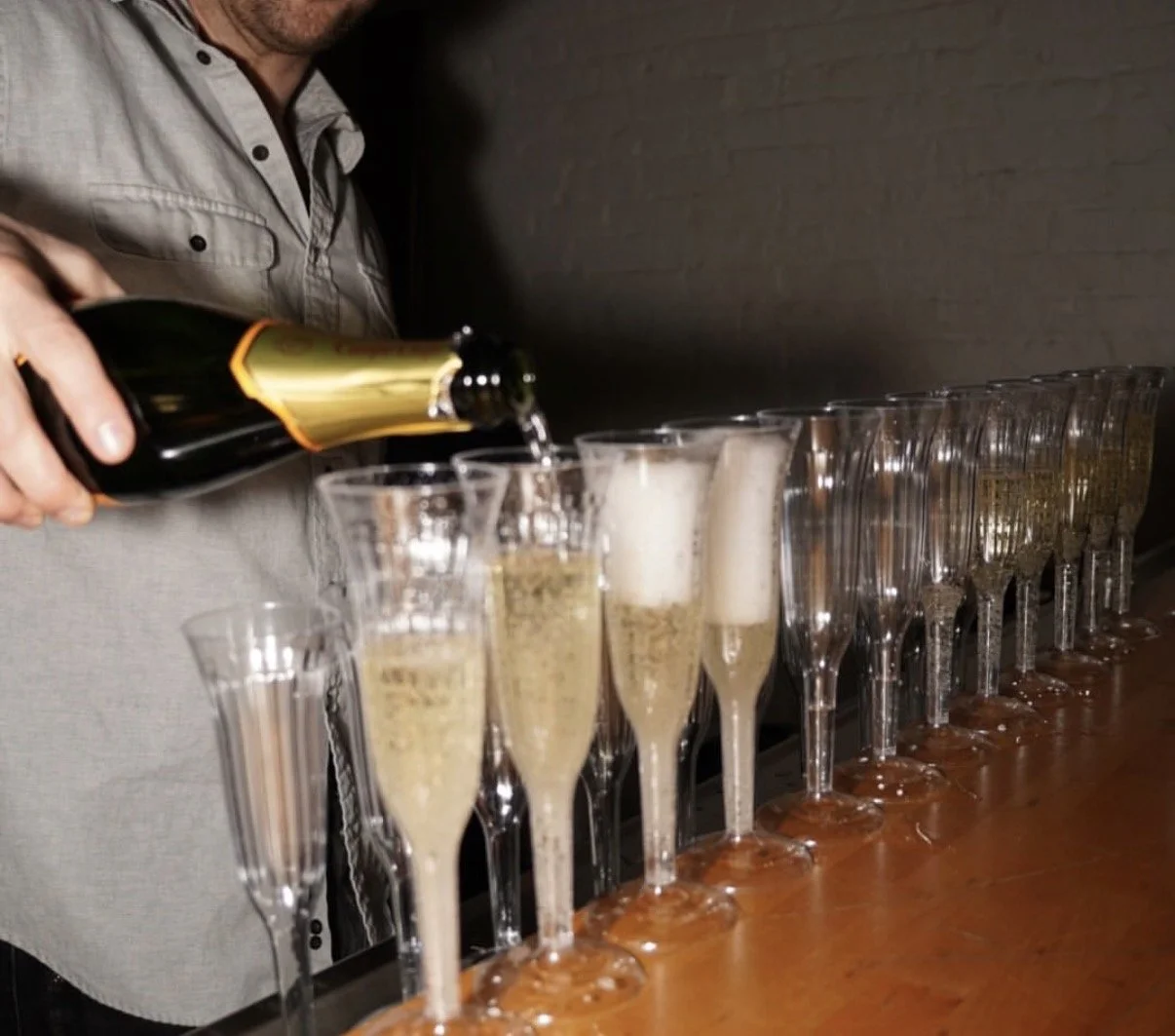 Person pouring champagne into a row of flutes on a bar counter, with foam forming at the top of each glass.
