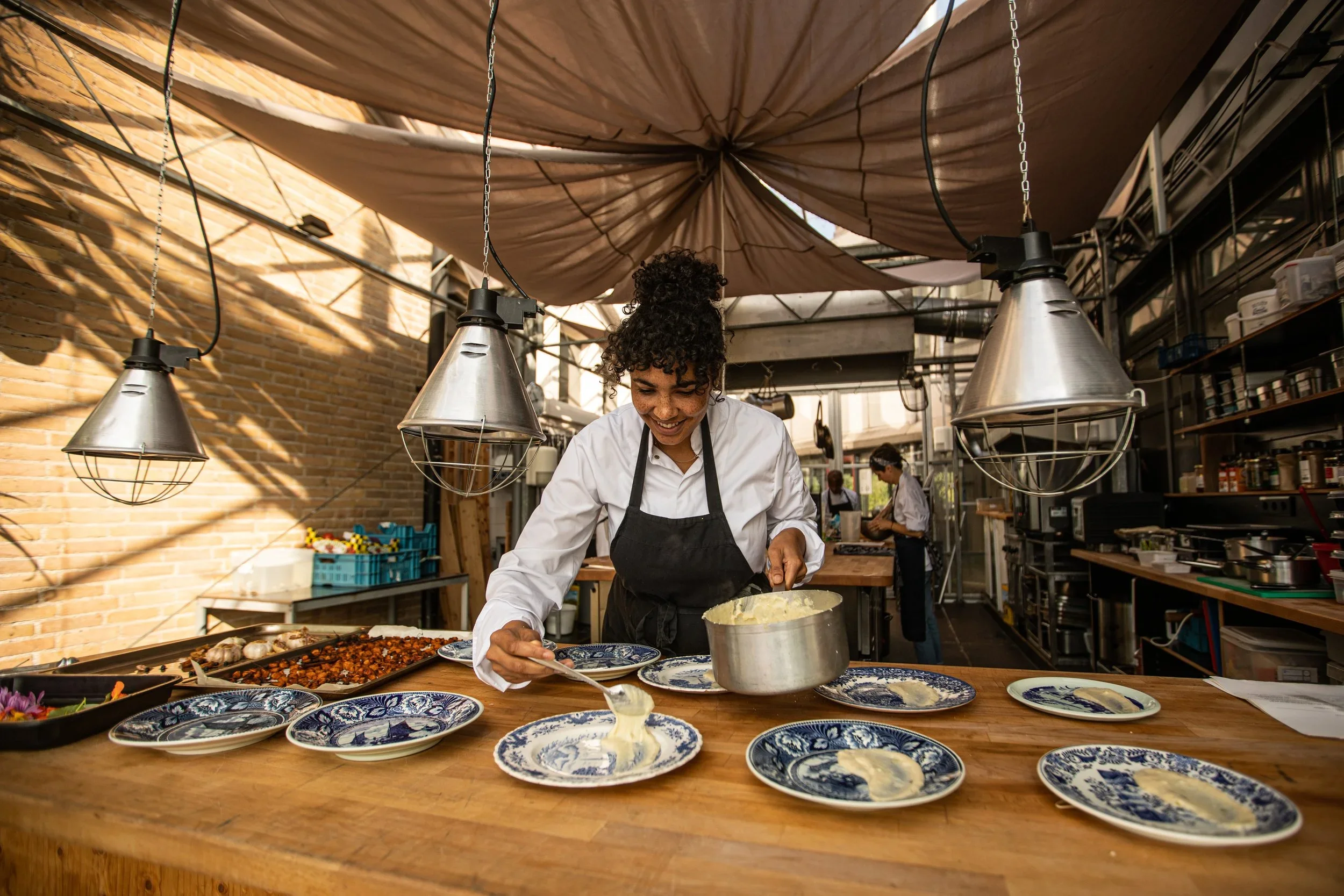 A chef serving mashed potatoes onto decorative blue and white plates in a rustic, industrial-style kitchen with exposed brick walls and hanging pendant lights.