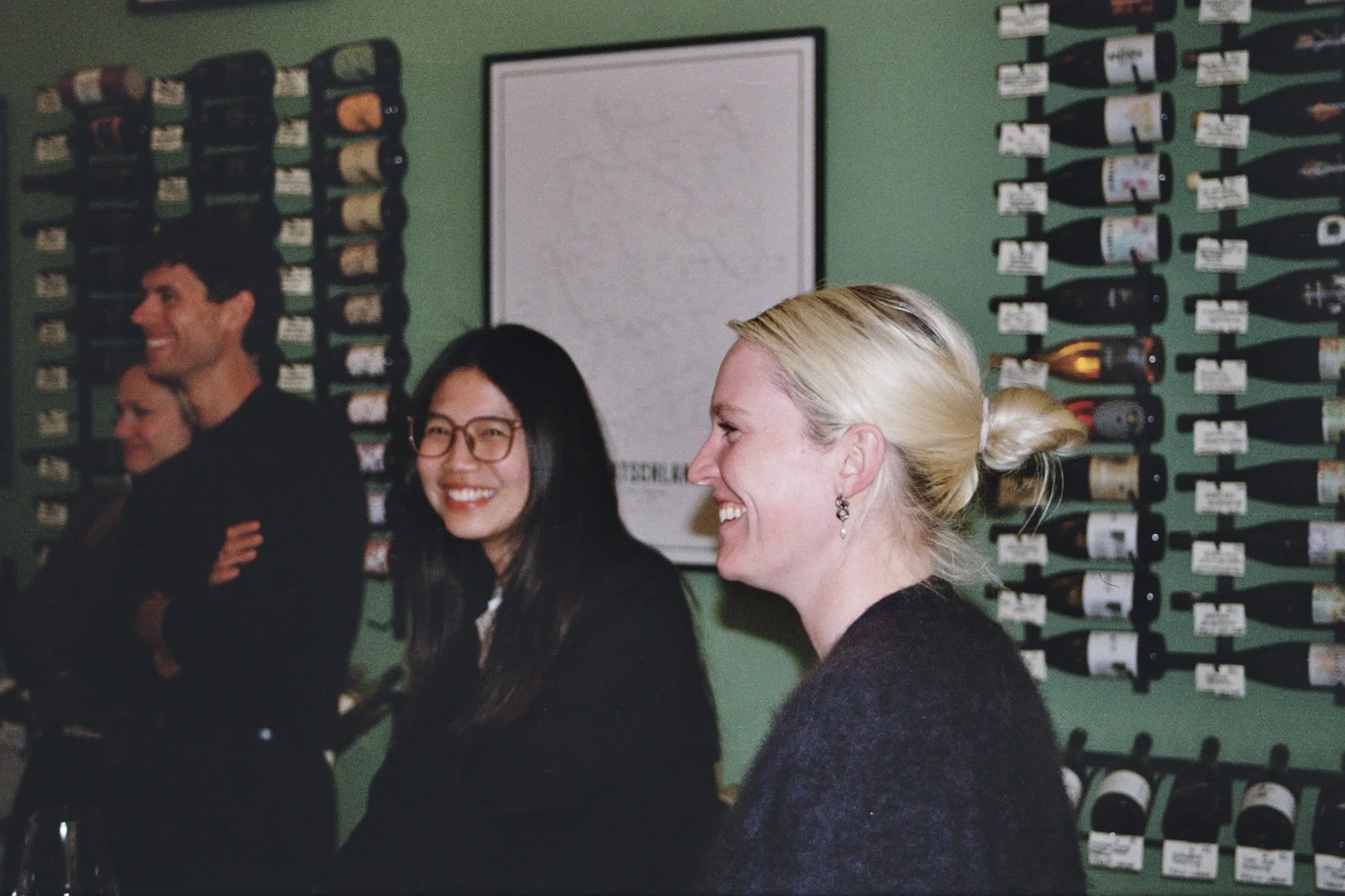 Group of people smiling and laughing at a wine tasting event with rows of wine bottles on the green wall behind them.